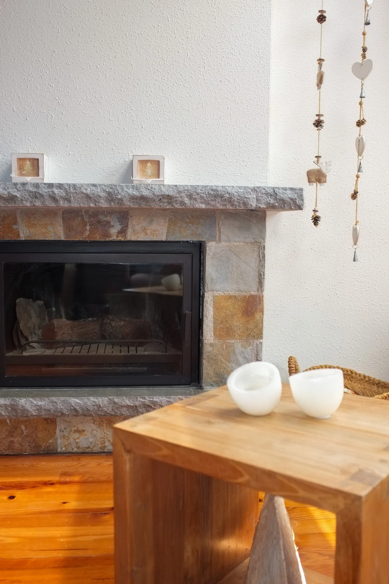 Hoguera de leña en una chimenea de piedra en una sala de estar, con decoración de árbol de Navidad enmarcada en la pared y jarrones blancos en una mesa de madera.