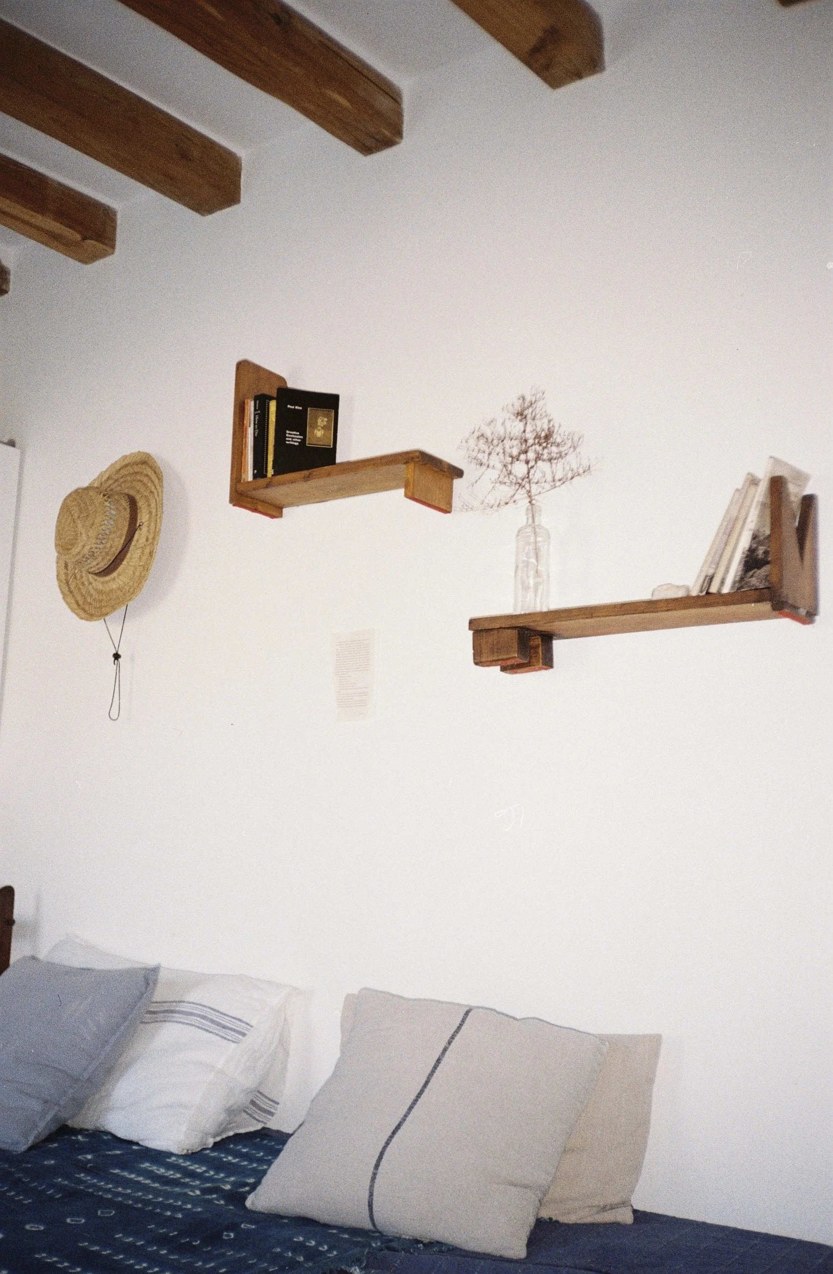 Cama con varias almohadas, pared blanca con estantes de madera con libros y una rama seca en un frasco de vidrio, sombrero de paja colgado en la pared.