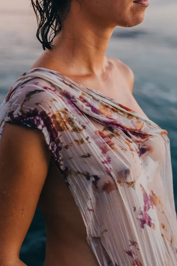Mujer con cabello húmedo, usando blusa de colores claros y estampados florales, en un entorno con agua al fondo.