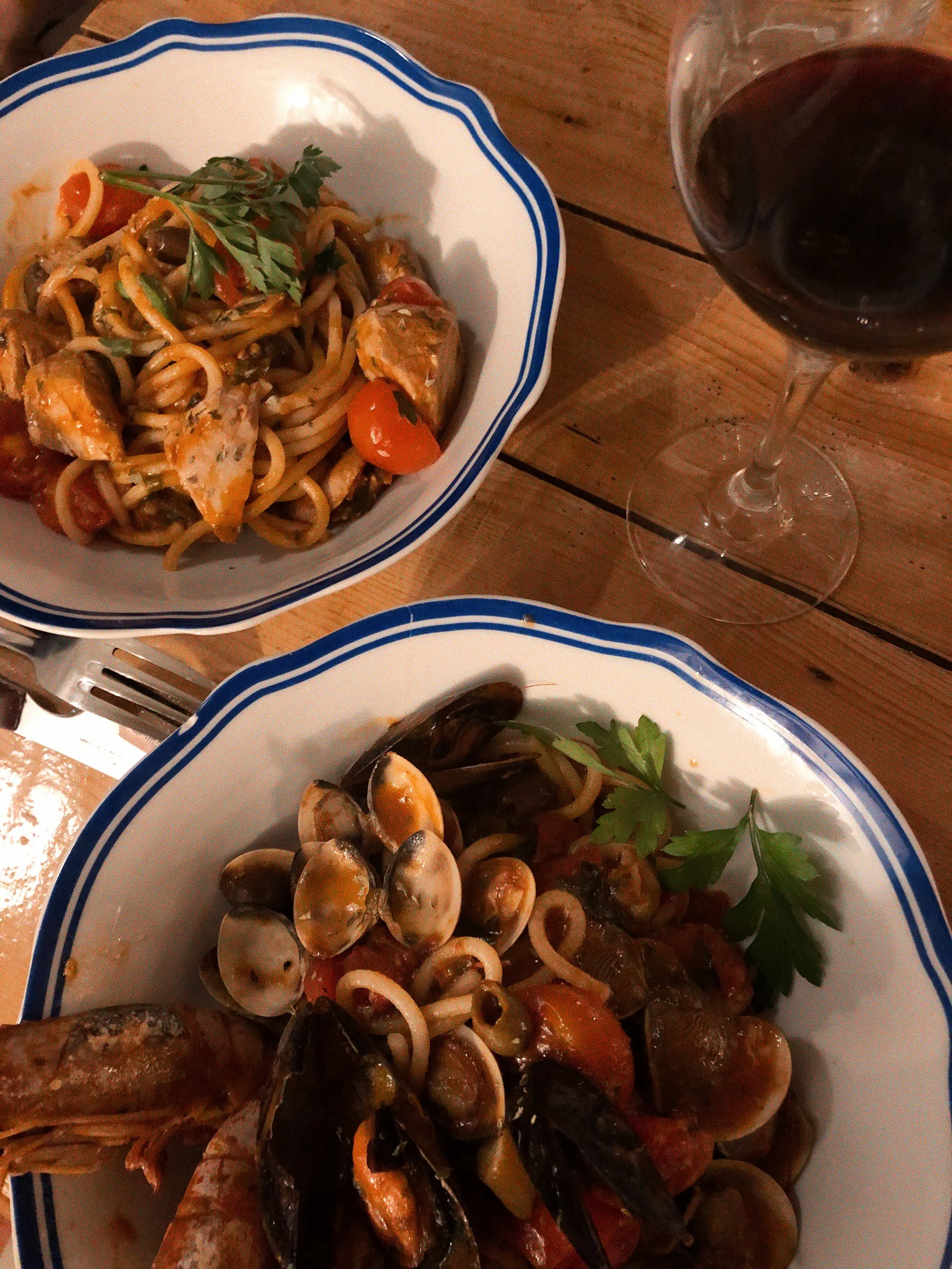 Platos de pasta con mariscos y sopa con mariscos, acompañados de una copa de vino tinto sobre una mesa de madera.