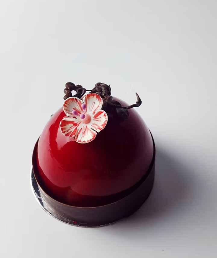 Pastel de mousse en forma de esfera cubierta con glaseado rojo, decorado con una flor y pequeñas figuras de chocolate.
