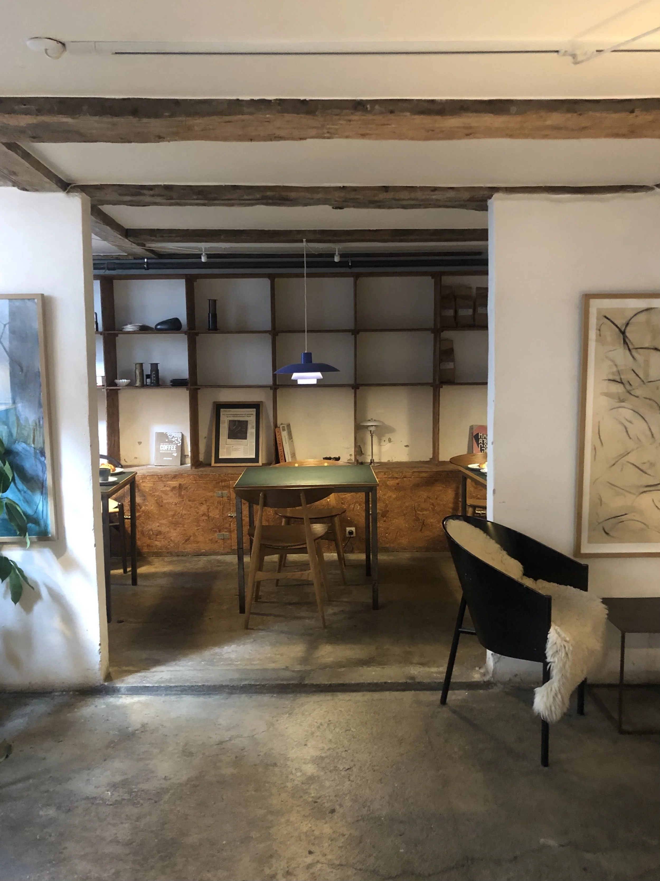 Interior de un espacio con mesas y sillas de madera, estantes de madera en la pared de fondo, lámpara colgante azul y decoración moderna.