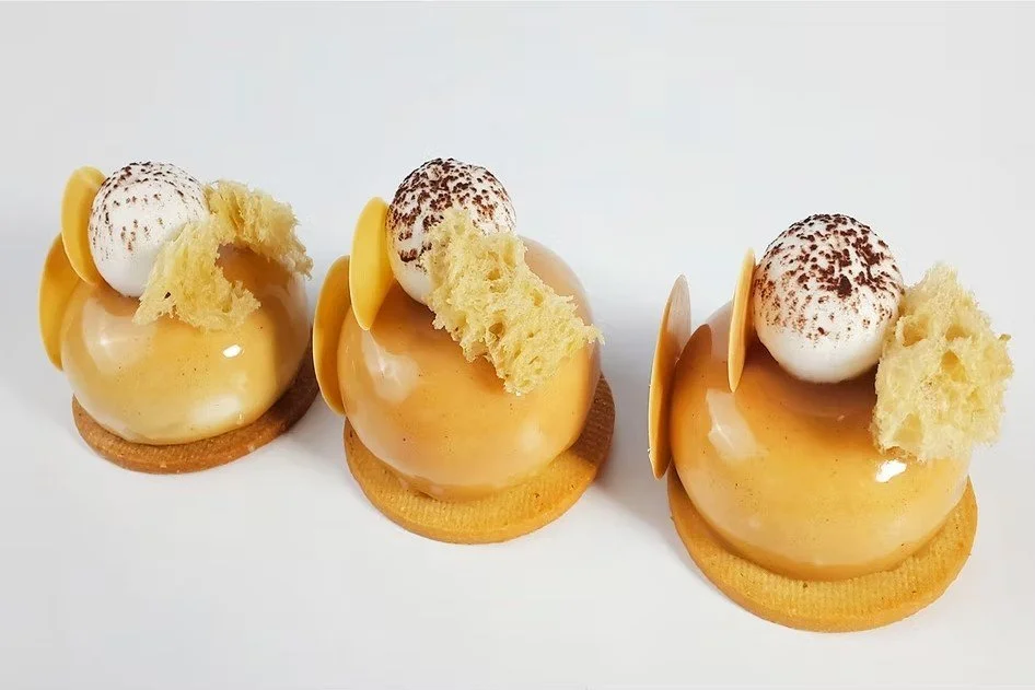 Tres postres con forma de tarta, glaseados de color caramelo, decorados con gotas de chocolate, merengue y galletas, cada uno sobre una base redonda.