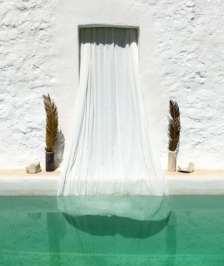 Cortina de tela blanca en una pared blanca que fluye sobre una piscina de agua turquesa, con dos plantas secas en macetas y algunas rocas decorativas.