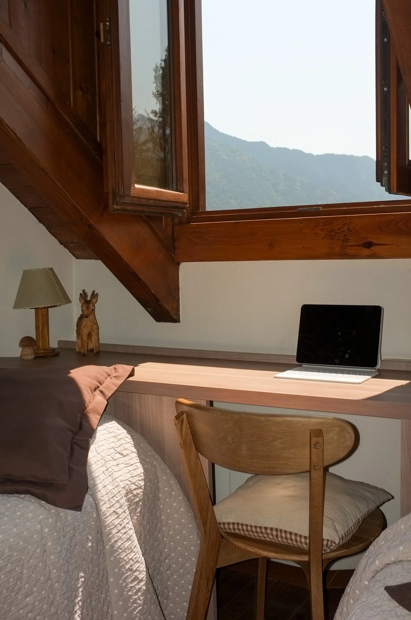 Vista de una habitación con ventanas abiertas, vista a montañas y vegetación, escritorio con computadora portátil, lámpara, figura de un ciervo de peluche, y cama con cobijas, en un ambiente con madera.