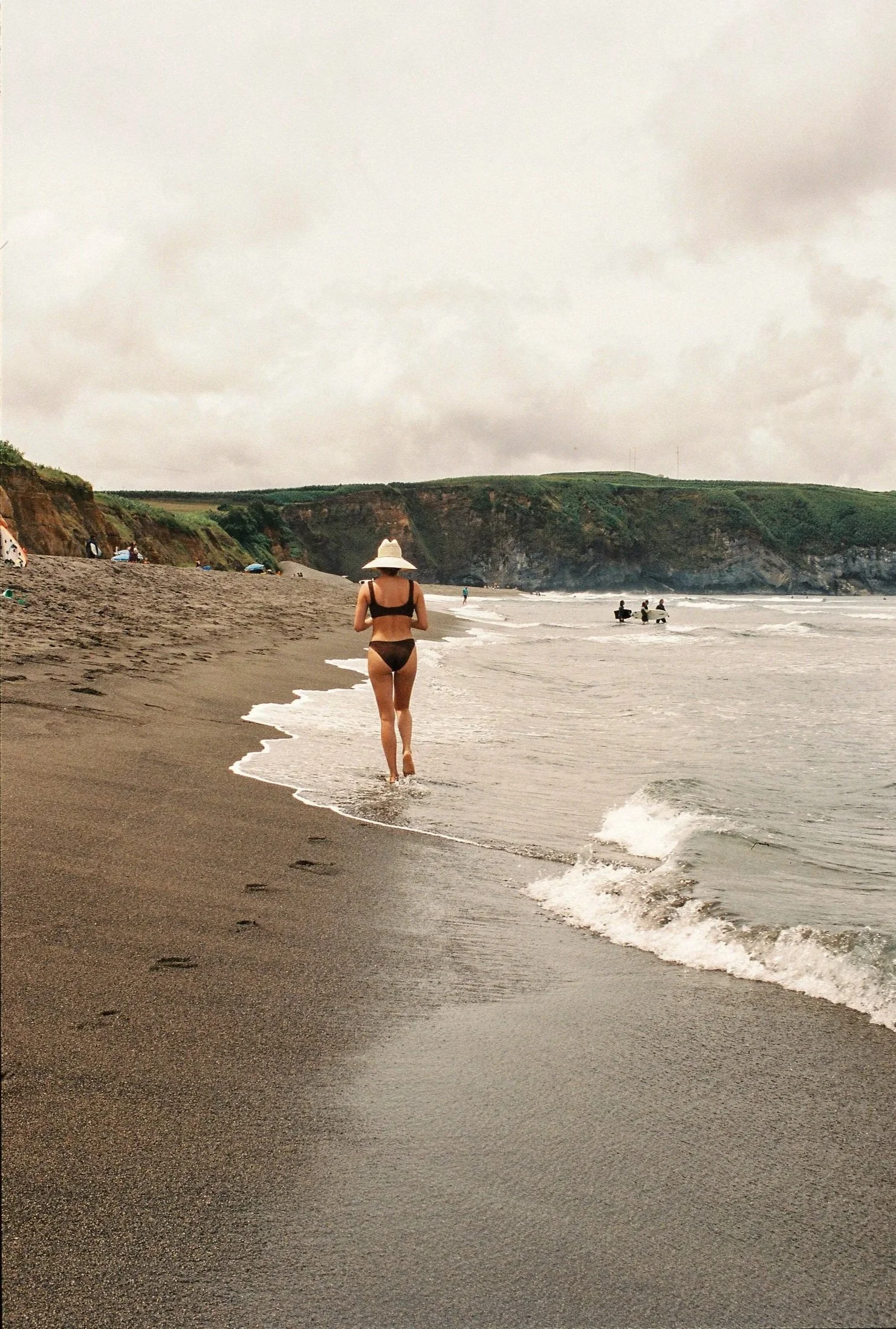 Mujer con traje de baño negro y sombrero caminando por la orilla de una playa con arena oscura, olas y acantilados verdes en el fondo, cielo nublado.
