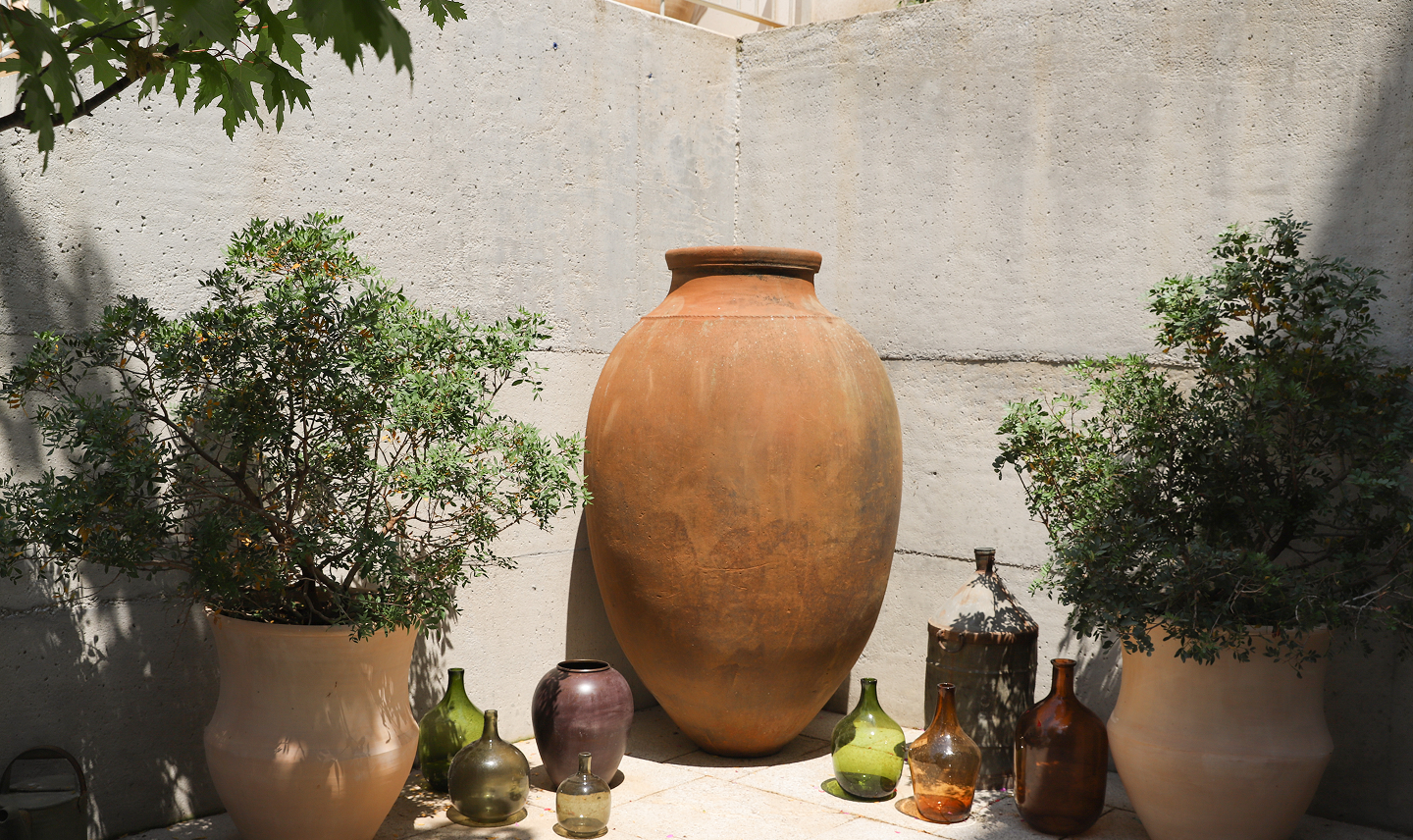 Jarrón grande de barro en el centro con varias vasijas de diferentes tamaños y colores a su alrededor, plantas a la izquierda y derecha, muro de concreto de fondo.