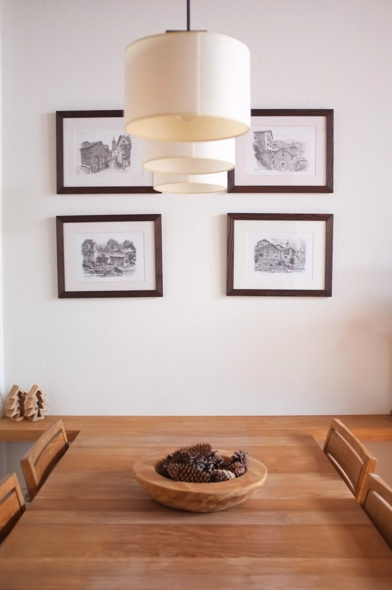Mesa de madera con centro de madera con piñas decorativas, paredes blancas con cuatro cuadros enmarcados con escenas de casas antiguas, lámpara blanca colgante, sillas de madera, decoraciones de pequeños árboles en la esquina izquierda.