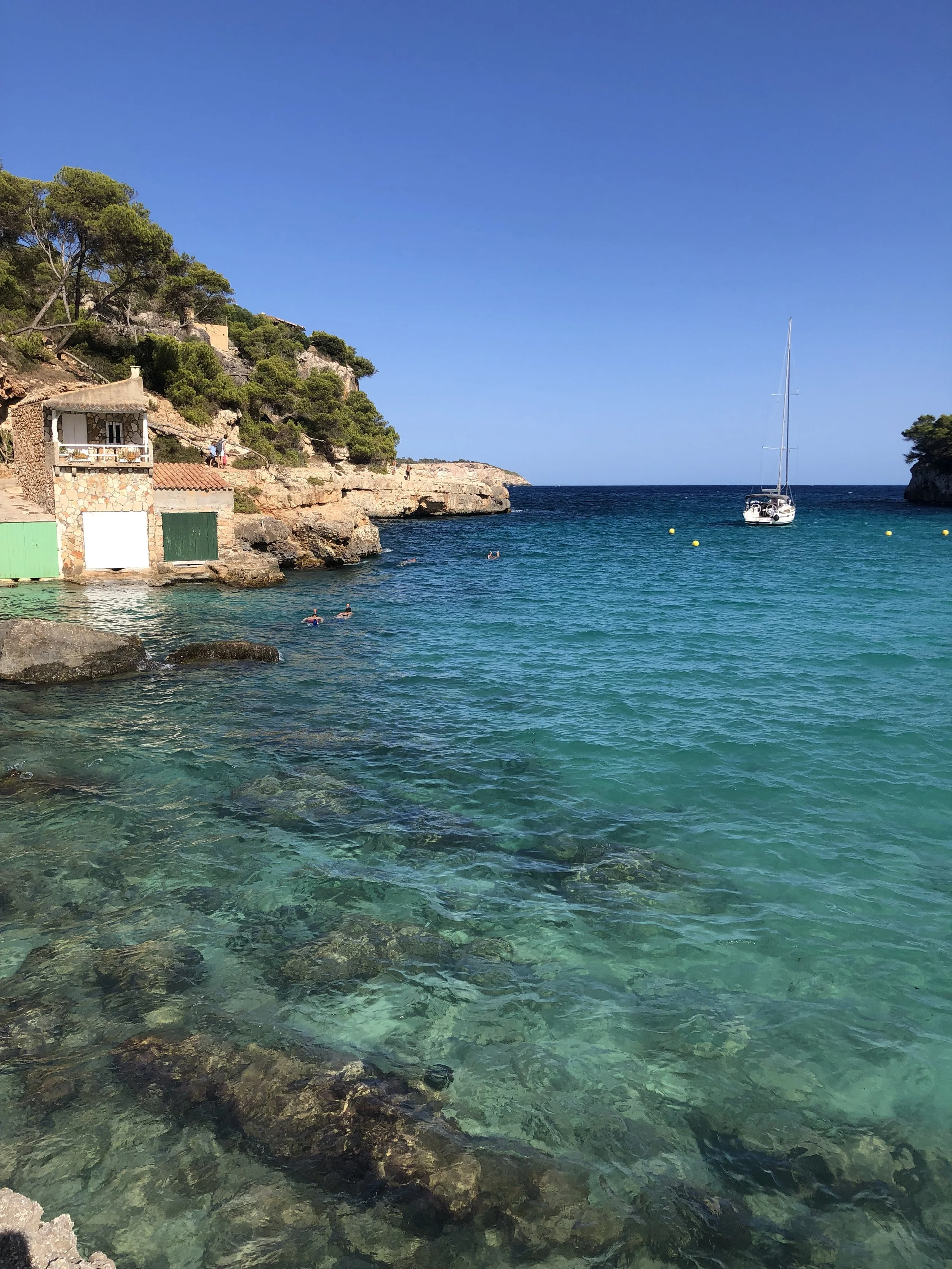 Vista de una playa con agua clara, rocas en la orilla, un barco y un acantilado con vegetación y casas pequeñas.