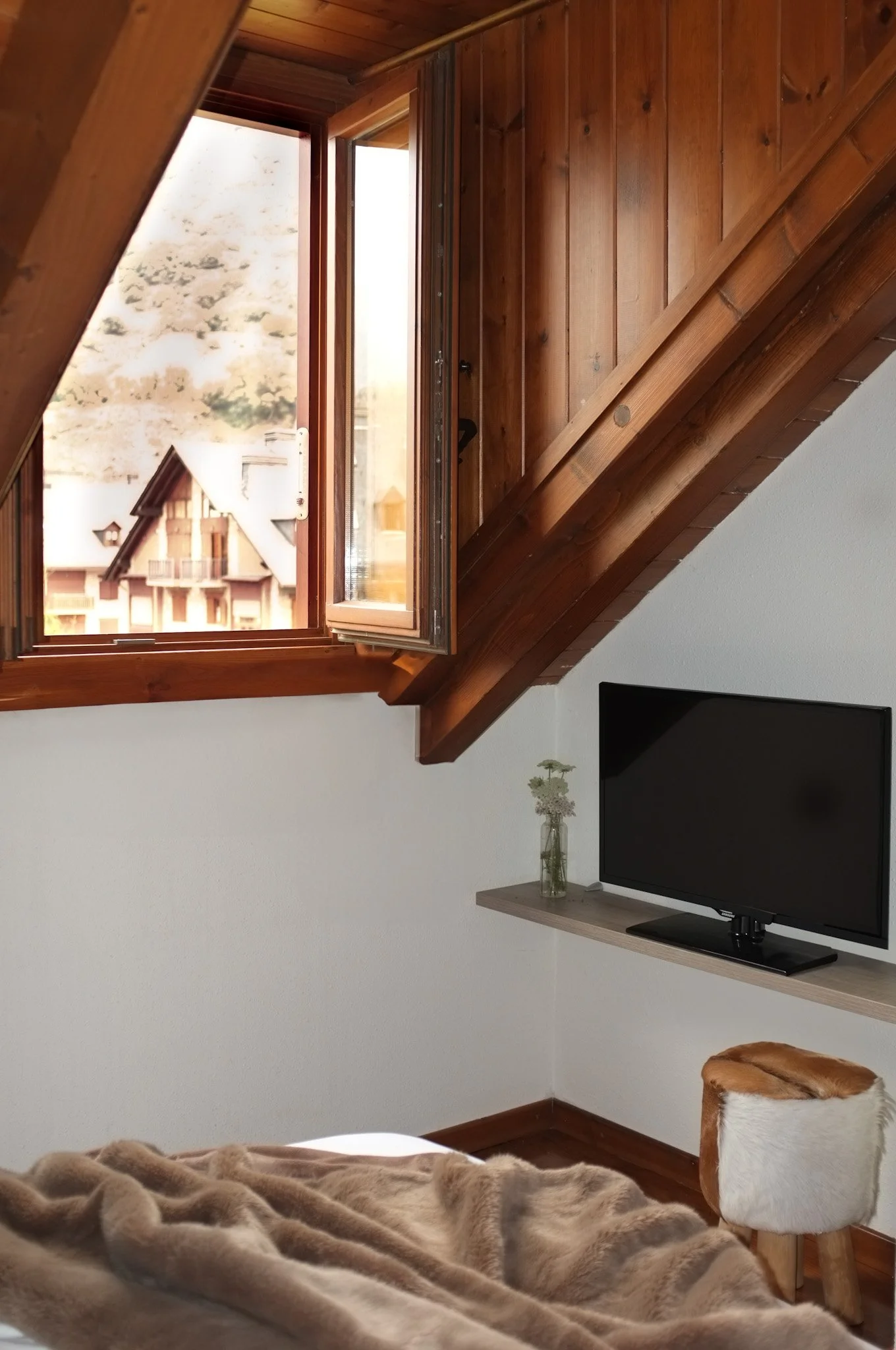 Habitación con pared blanca, con ventana de madera que da vista a casas y montañas, televisor en una repisa con flores en un jarrón, y una silla con cubierta de peluche beige.