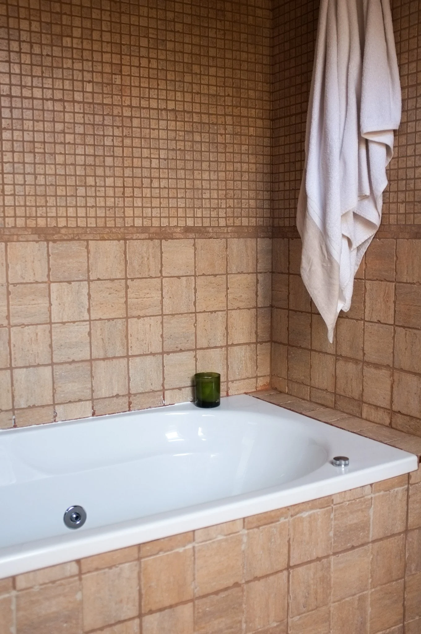 Una tina de baño blanca, una vela verde y una toalla blanca colgada en la pared, paredes de azulejos marrones en un baño.