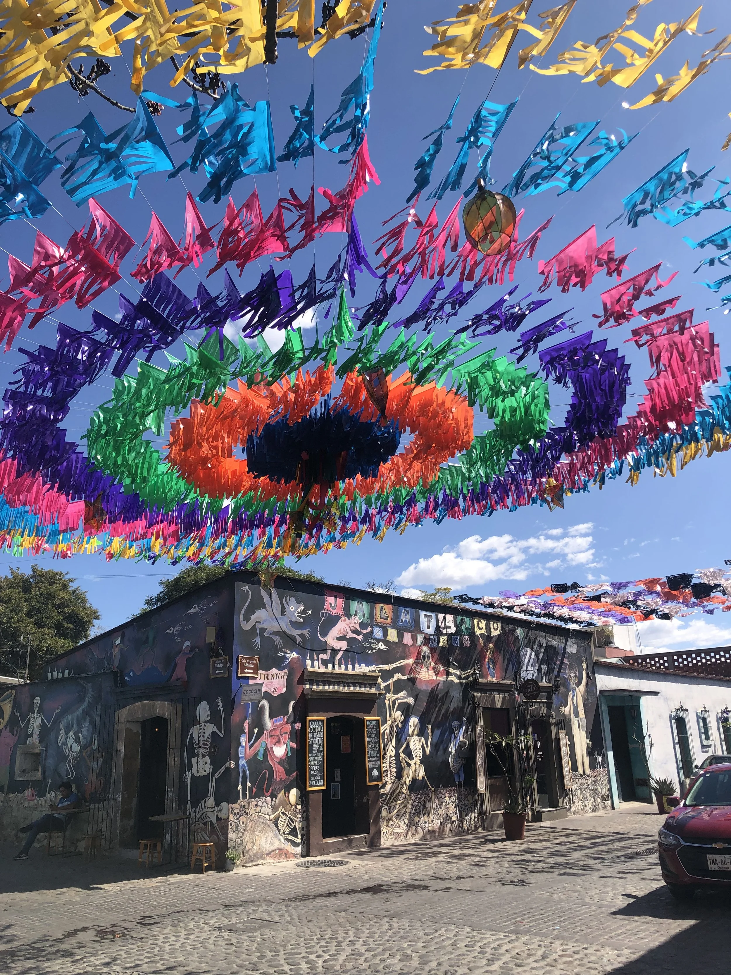 Tienda decorada con arte mural con esqueletos y criaturas fantásticas, con banderines de colores sobre la calle, cielo despejado y un coche rojo estacionado.
