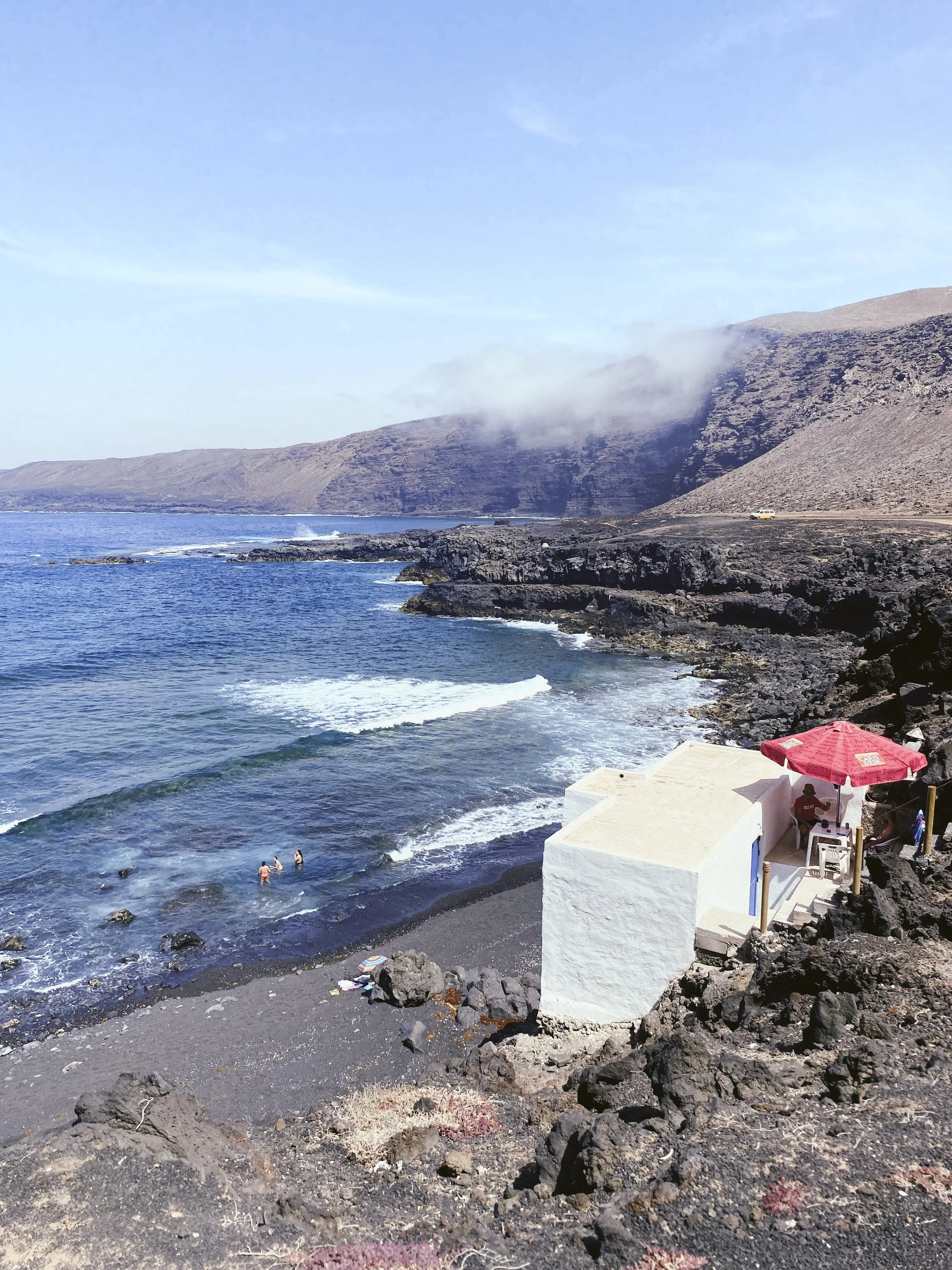 Costa rocosa con playa de arena oscura, olas suaves, y un restaurante con sombrilla roja y sombra en el lado derecho. En el mar, personas disfrutando el agua y montañas en el fondo con algunas nubes.