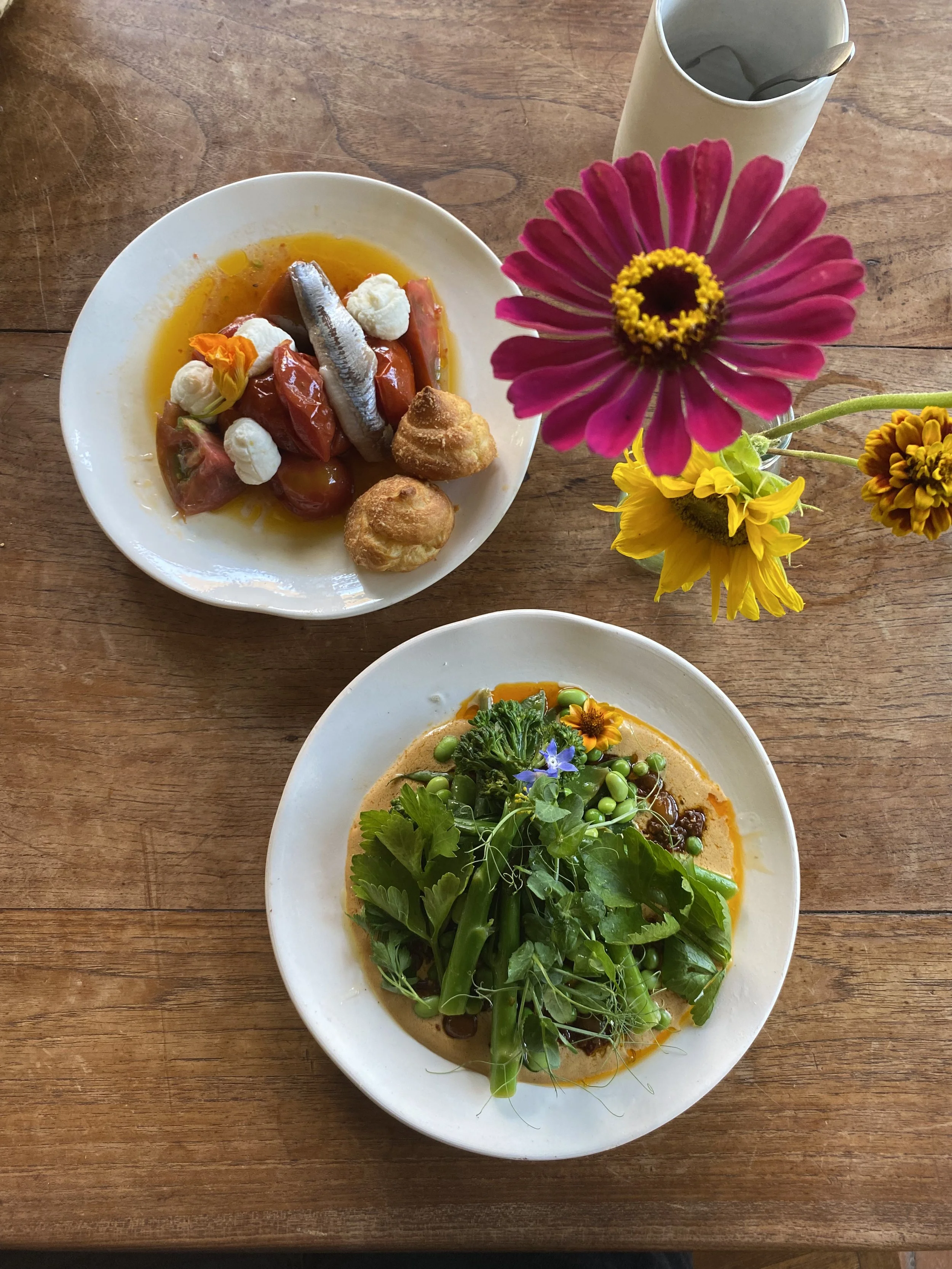 Plato con tomates, sardina, y pequeños bocadillos en un plato, y otro plato con verduras frescas y flores comestibles, en una mesa de madera, acompañados de un florero con flores de colores vivos.