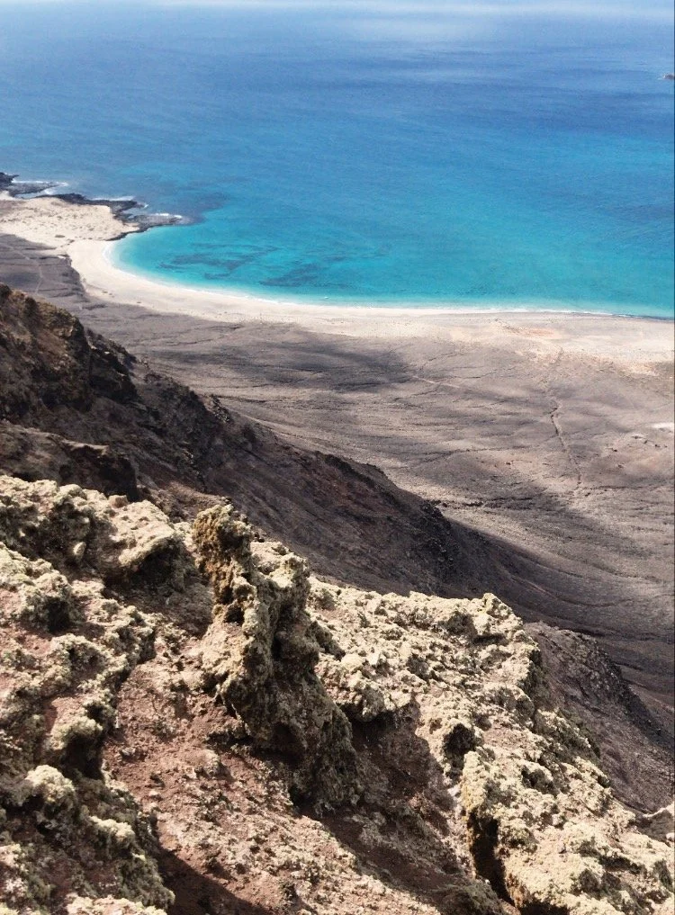 Vista desde una montaña mostrando un mar azul y una playa con rocas en la orilla.