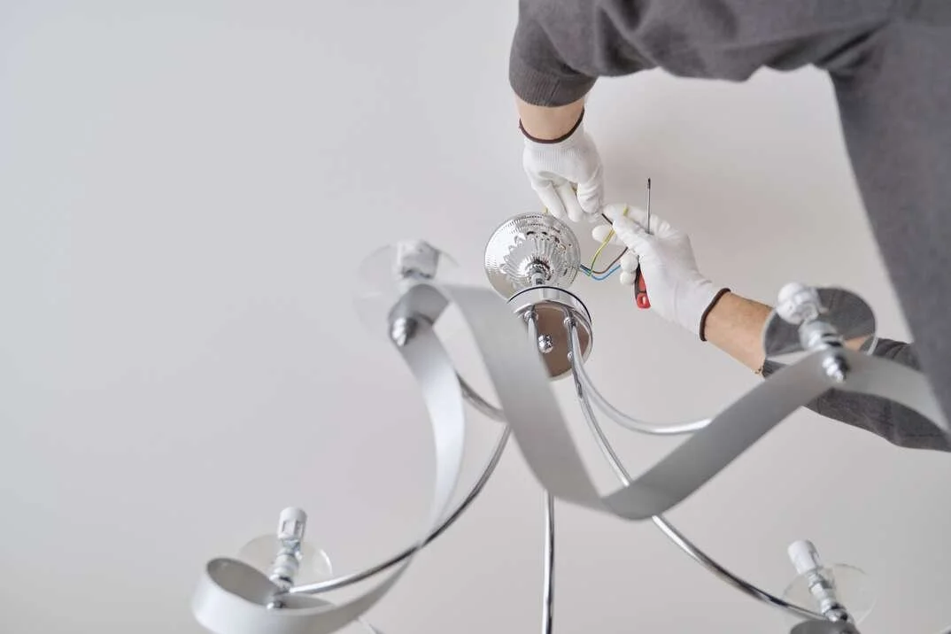 A person wearing white gloves installing or repairing a modern chandelier with metallic arms and light sockets.