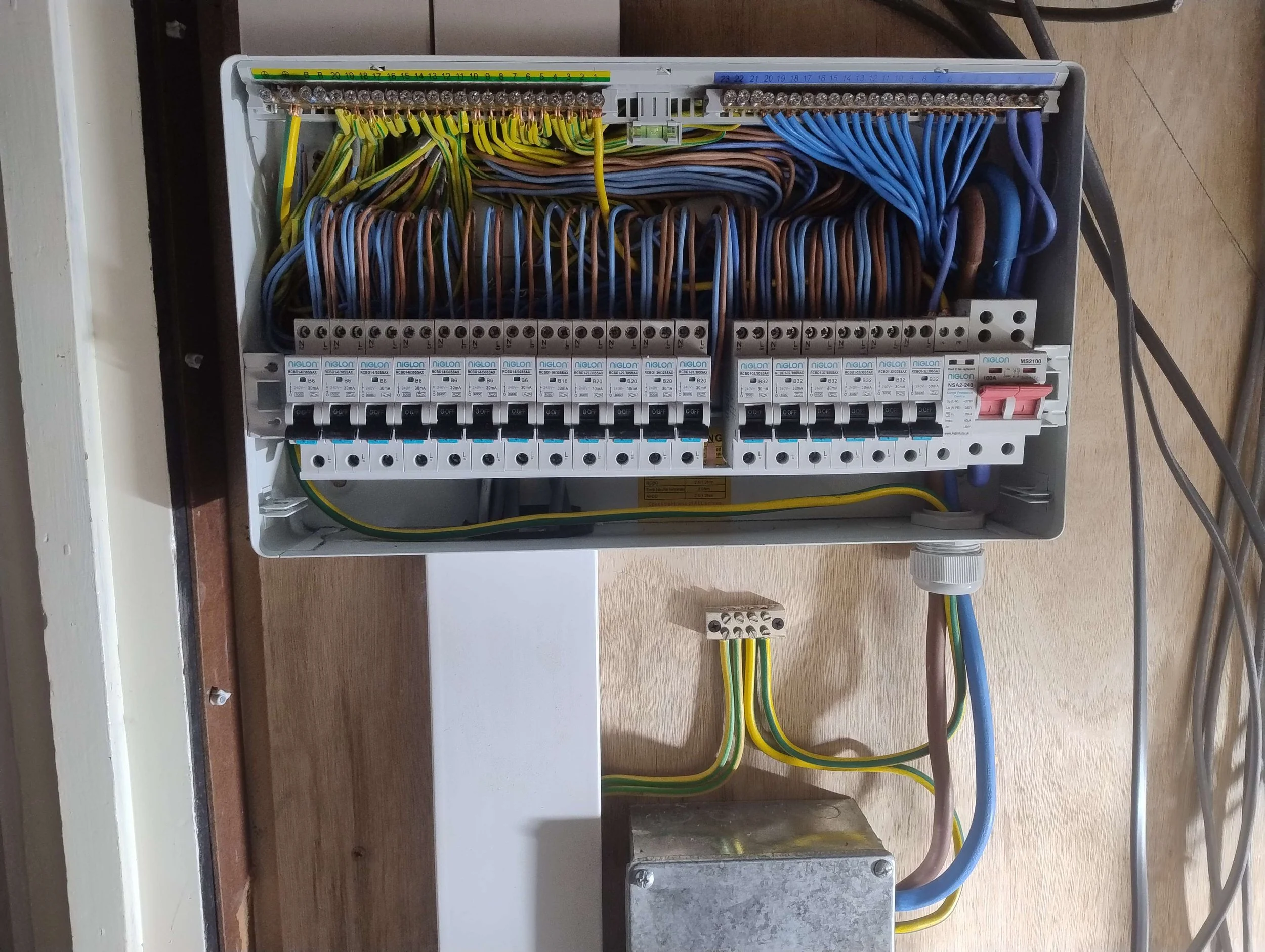 Open electrical panel with multiple colored wires connected to circuit breakers and switches, mounted on a wooden surface.