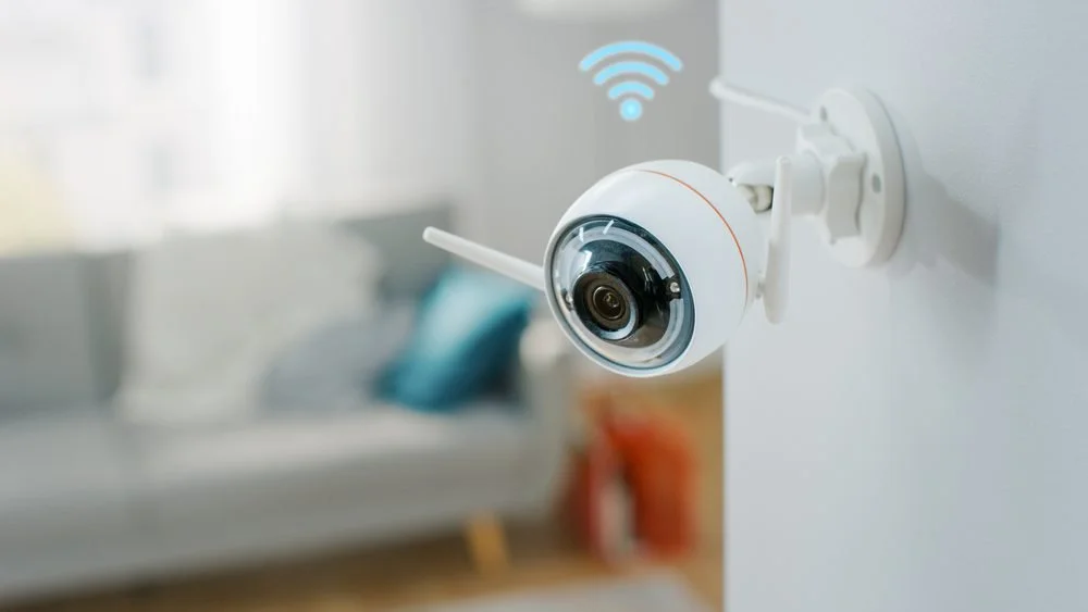 Wireless security camera mounted on a wall inside a home, with a blurred living room in the background.