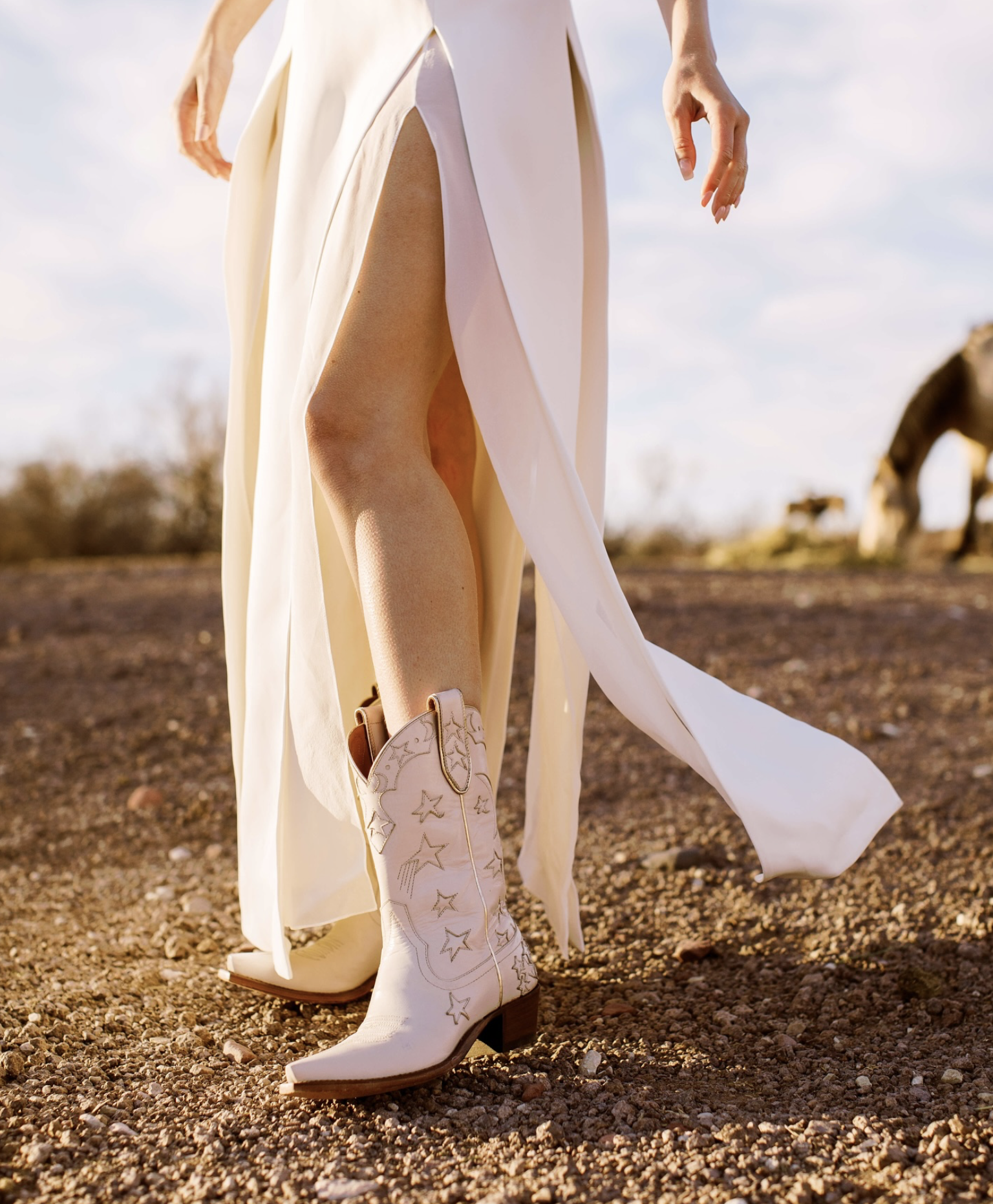 Close-up of a person wearing white cowboy boots with star and moon designs, a flowing cream-colored dress with high slit, standing on a rocky terrain during sunset.