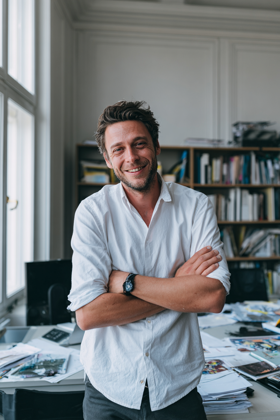 A smiling man with dark hair and a beard, wearing a white button-up shirt with sleeves rolled up, stands in an office with arms crossed. The office has large windows, a cluttered desk with papers, and a bookshelf filled with books in the background.