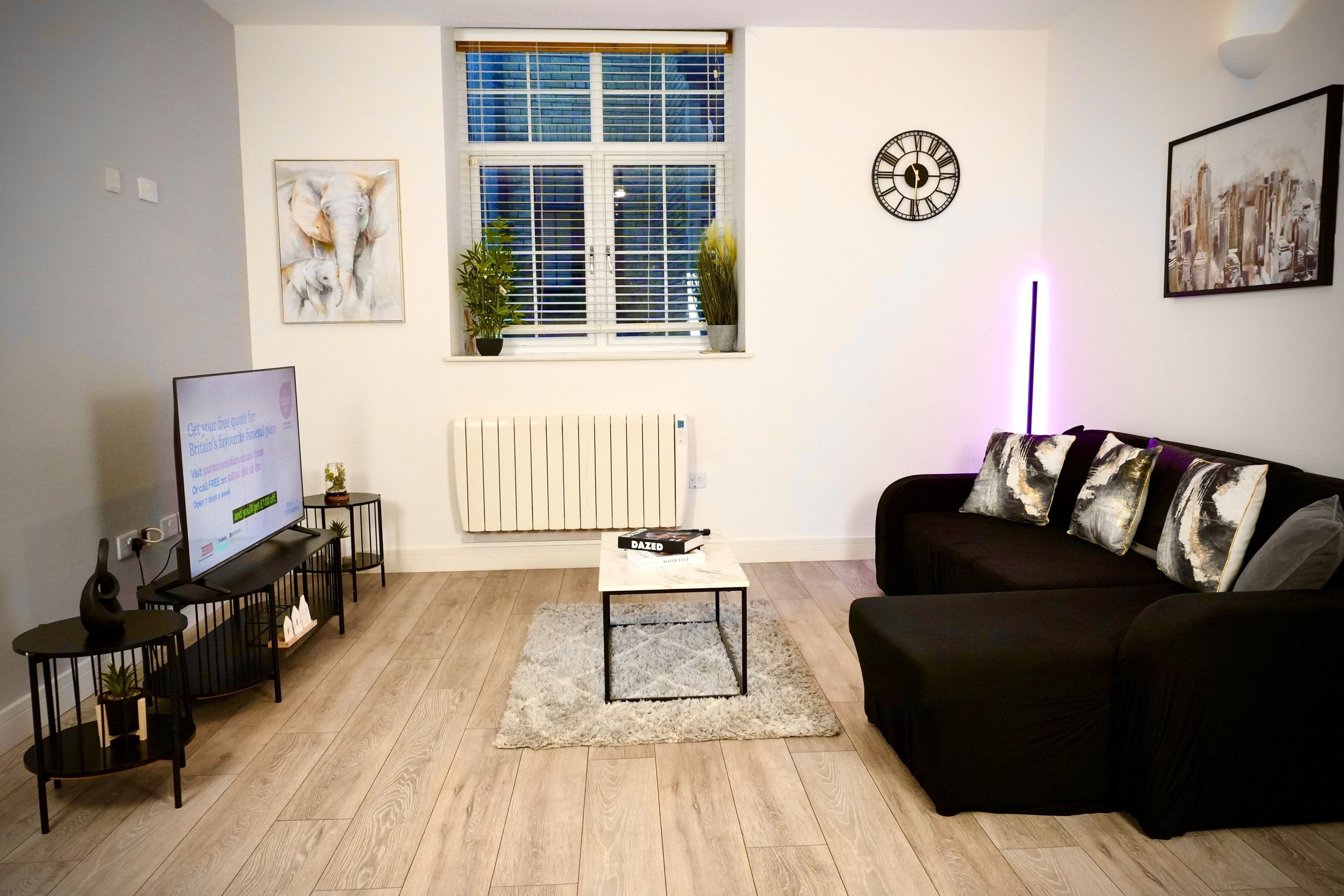 Modern living room with a black sofa, patterned cushions, a small coffee table on a beige rug, a wall-mounted TV on a black stand, a window with white blinds, artwork on the walls, potted plants on the window sill, and a wall clock. The room has light wood flooring and white walls.