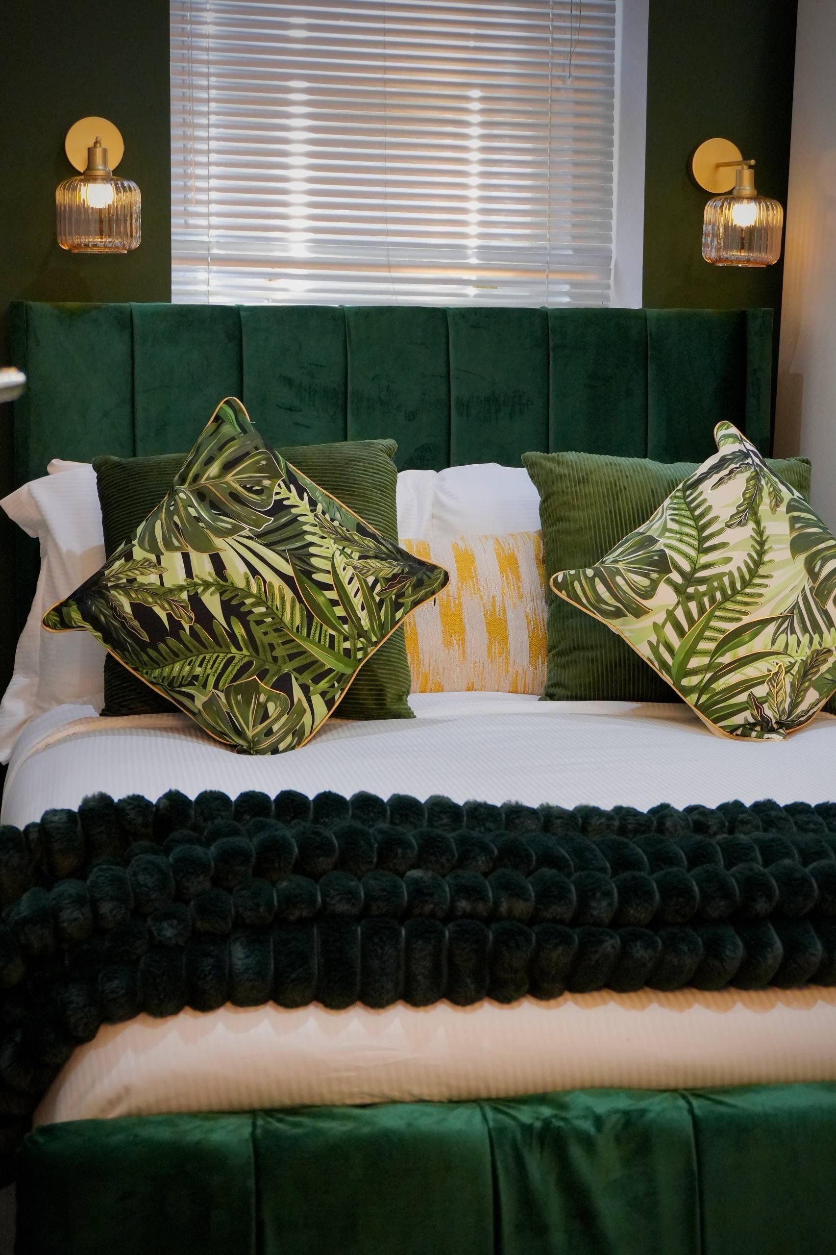 A hotel room with two white beds, one larger and one smaller, each with pillows and rolled towels with small bottles on top. A nightstand with a small lamp, a framed picture, and a plant sits between the beds. The room has plain white walls and a fitting wall-mounted light.