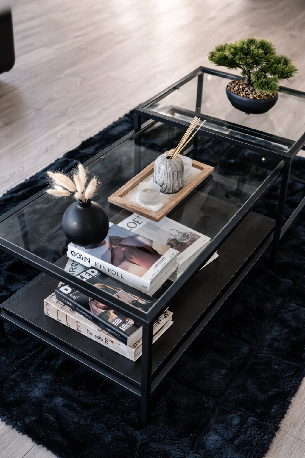 Modern coffee table with decorative items, magazines, a black vase with dried flowers, a tray with candles and reed diffuser, and a potted plant on top, all on a black shag rug.