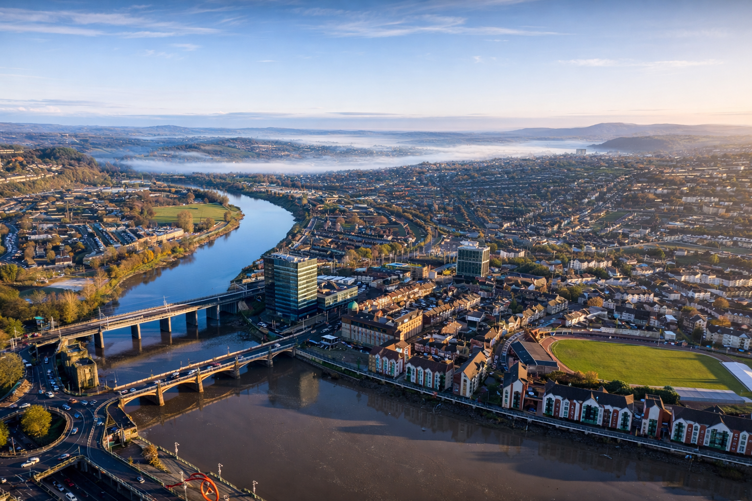 Aerial view of a cityscape with a river running through it, bridges crossing the river, and a mix of residential and commercial buildings, some tall, under clear skies.