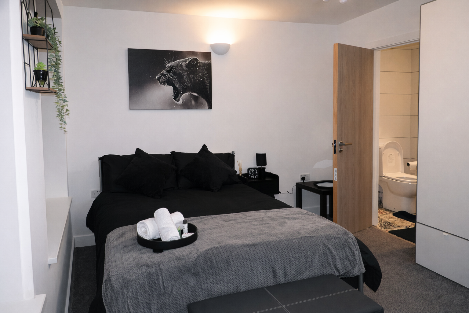 Modern bedroom with a black bed, black pillows, a gray blanket, and a tray with towels. The wall has a black and white lion artwork, and there is a small black nightstand with a lamp and decor. An open door reveals a bathroom with a toilet. A wardrobe is partially visible in the corner.