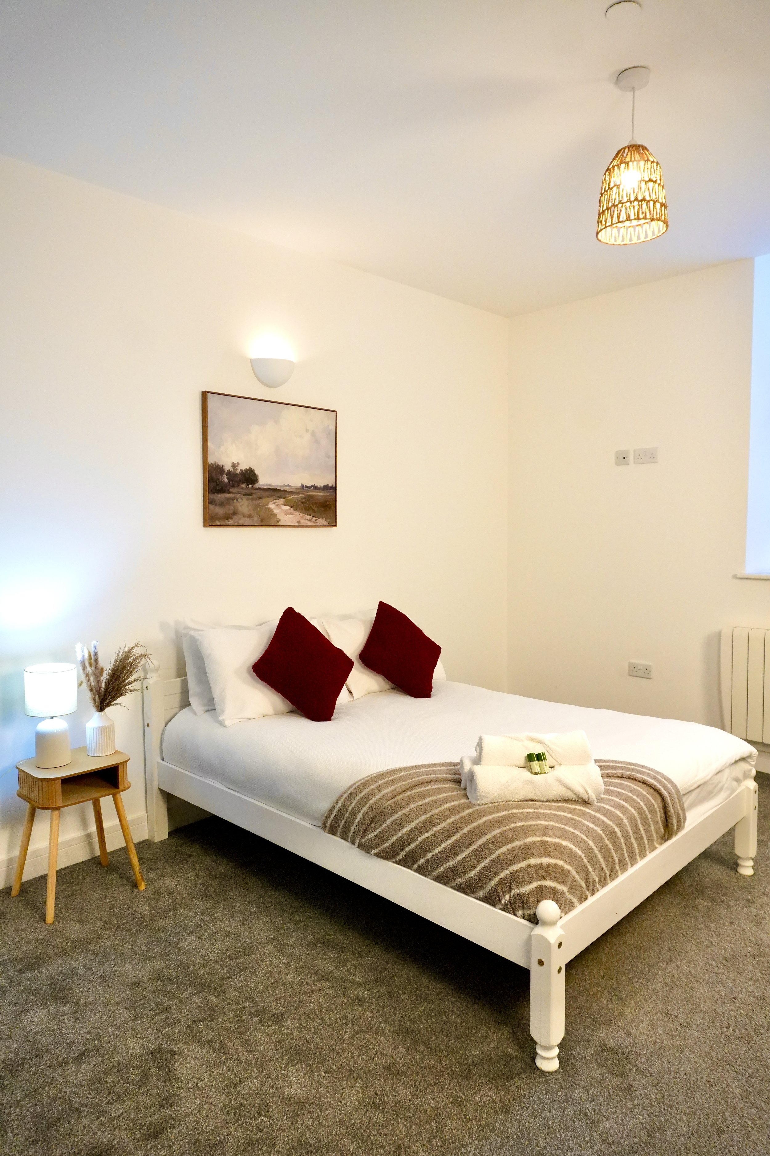 A neatly made bed with white linens, two red pillows, and a brown striped blanket on a white wooden bed frame in a minimalist bedroom. Beside the bed is a small wooden nightstand with a white lamp, a vase with dried grasses, and towels with toiletries. The room has white walls, gray carpet, a landscape painting, a wall-mounted light, and a woven ceiling light.