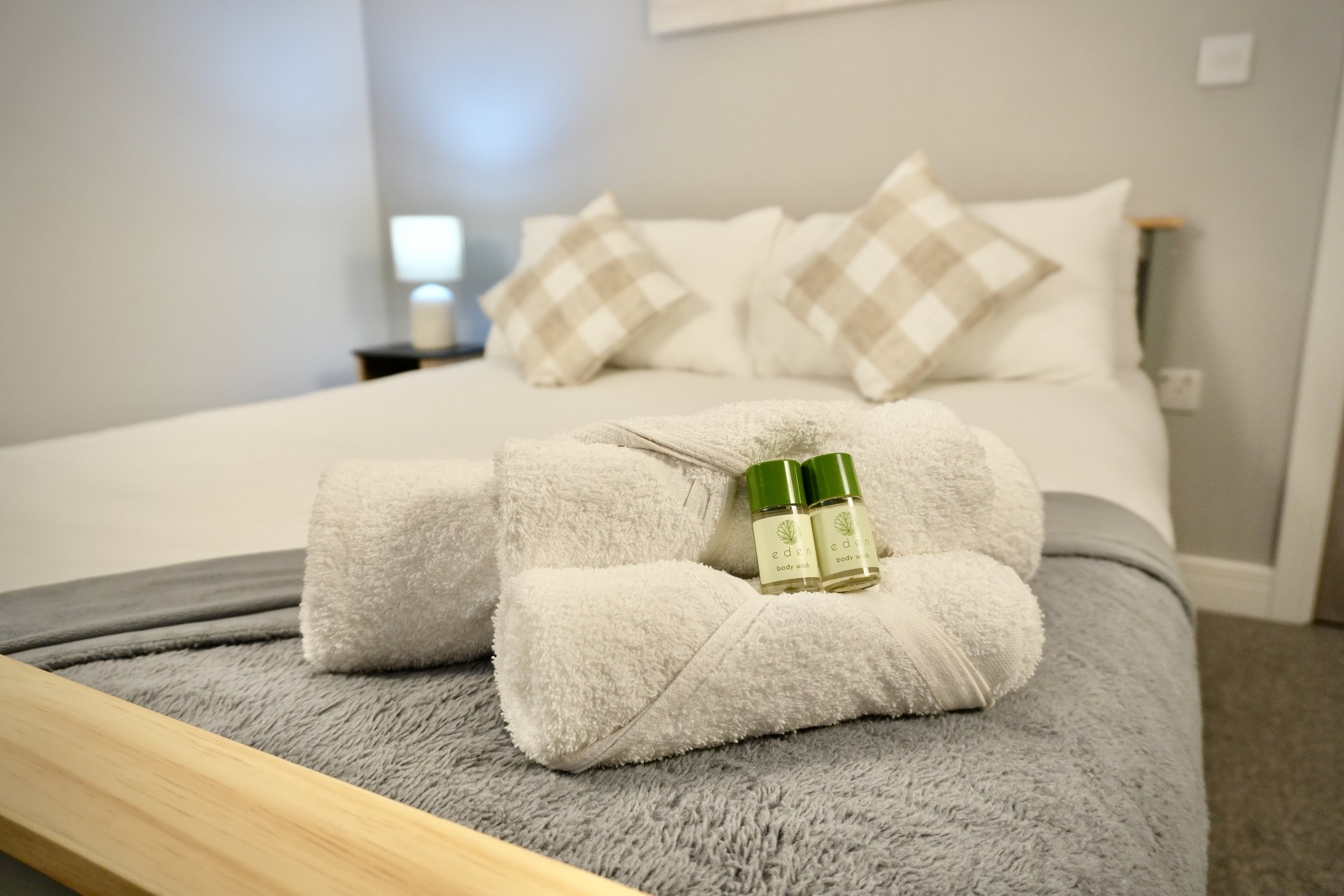 A neatly made hotel bed with white bedding and three checkered pillows. On the bed, two white towels are rolled with two small bottles of body wash placed on top.