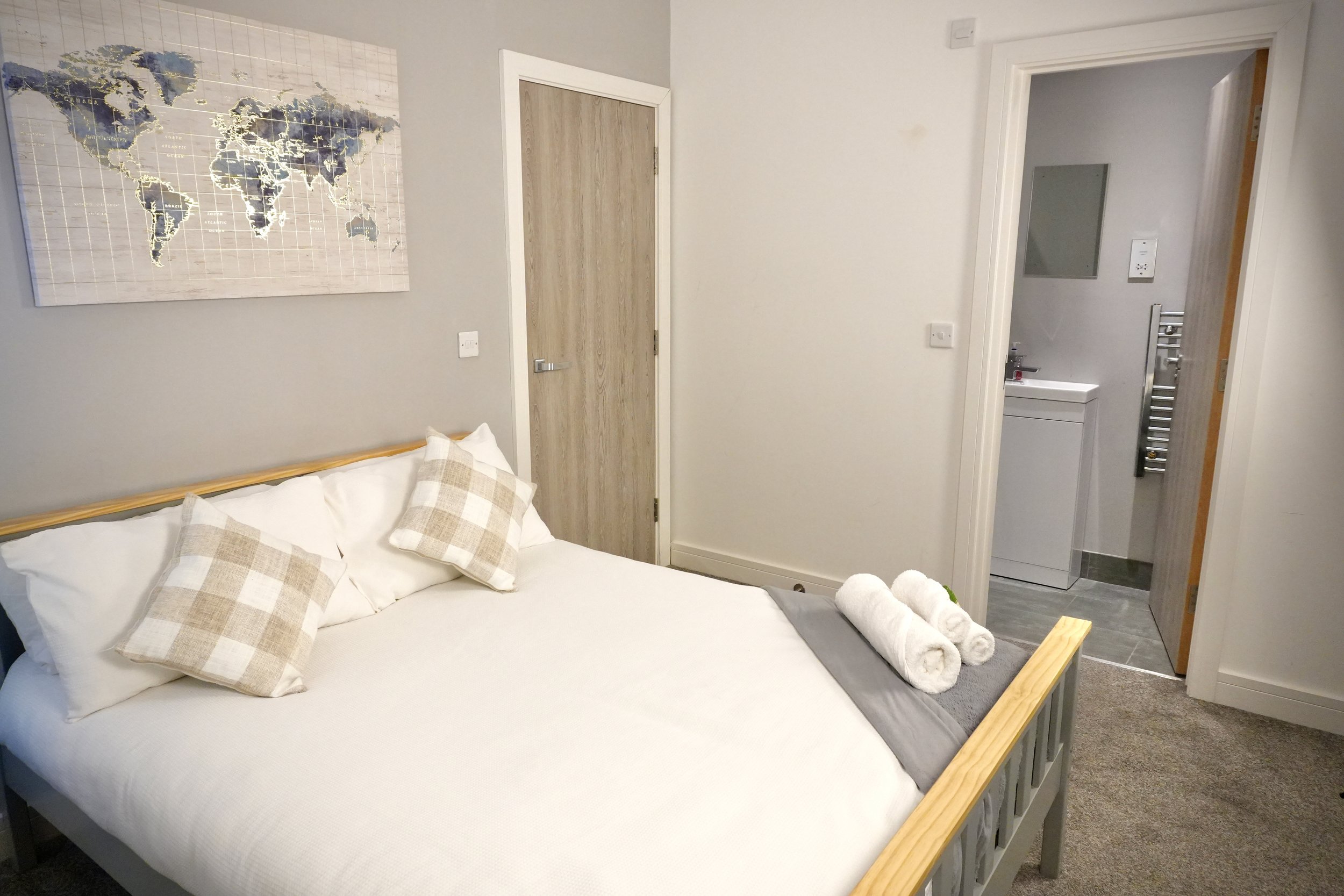 A bedroom with a bed, three checkered pillows, a world map wall art, and towels on the bed. An open door leads to a bathroom with a sink and mirror.