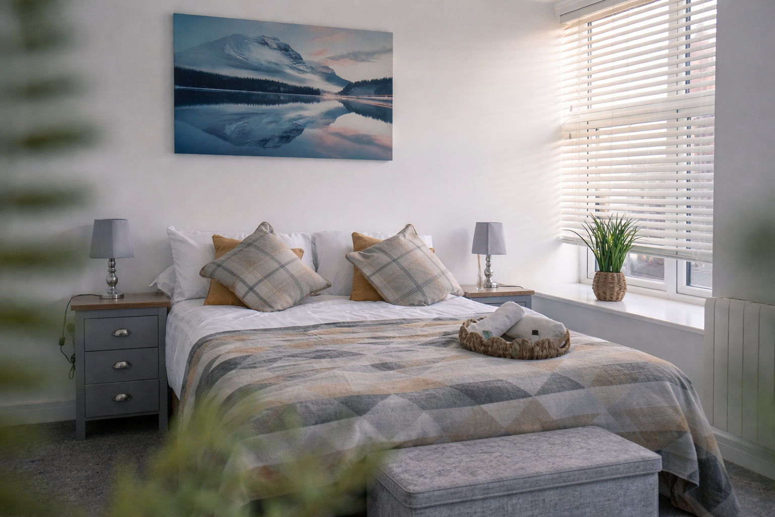 A cozy bedroom with a large bed, beige plaid pillows, and a quilted bedspread, flanked by matching bedside tables with lamps, a window with white blinds, a potted plant on the window sill, and a mountain landscape painting on the wall.