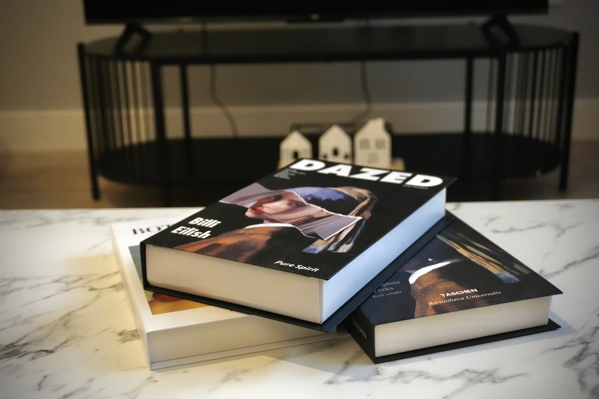 Two books stacked on a white marble table, with the top book titled 'Dazed' by Bill Elish, featuring a collage-style cover with a woman's face. The background includes a black metal bench and small decorative house models.