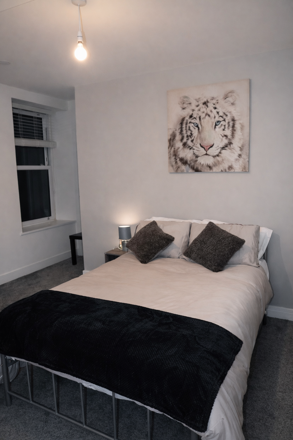 A bedroom with a bed, two pillows, a bedside table with a lamp, a painting of a white tiger on the wall, and a window with blinds. The room has gray walls and carpet, and a light bulb hanging from the ceiling.