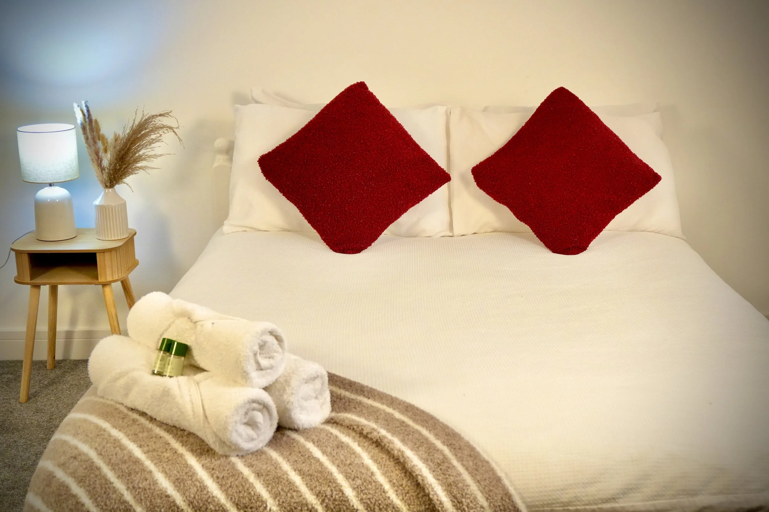A neatly made bedroom with a bed featuring white bedding, two red pillows, and a beige striped blanket. On the bed, there are three rolled white towels with small bottles on top. To the left, a wooden nightstand holds a white lamp and a vase with dried pampas grass. The wall behind is plain and light-colored.