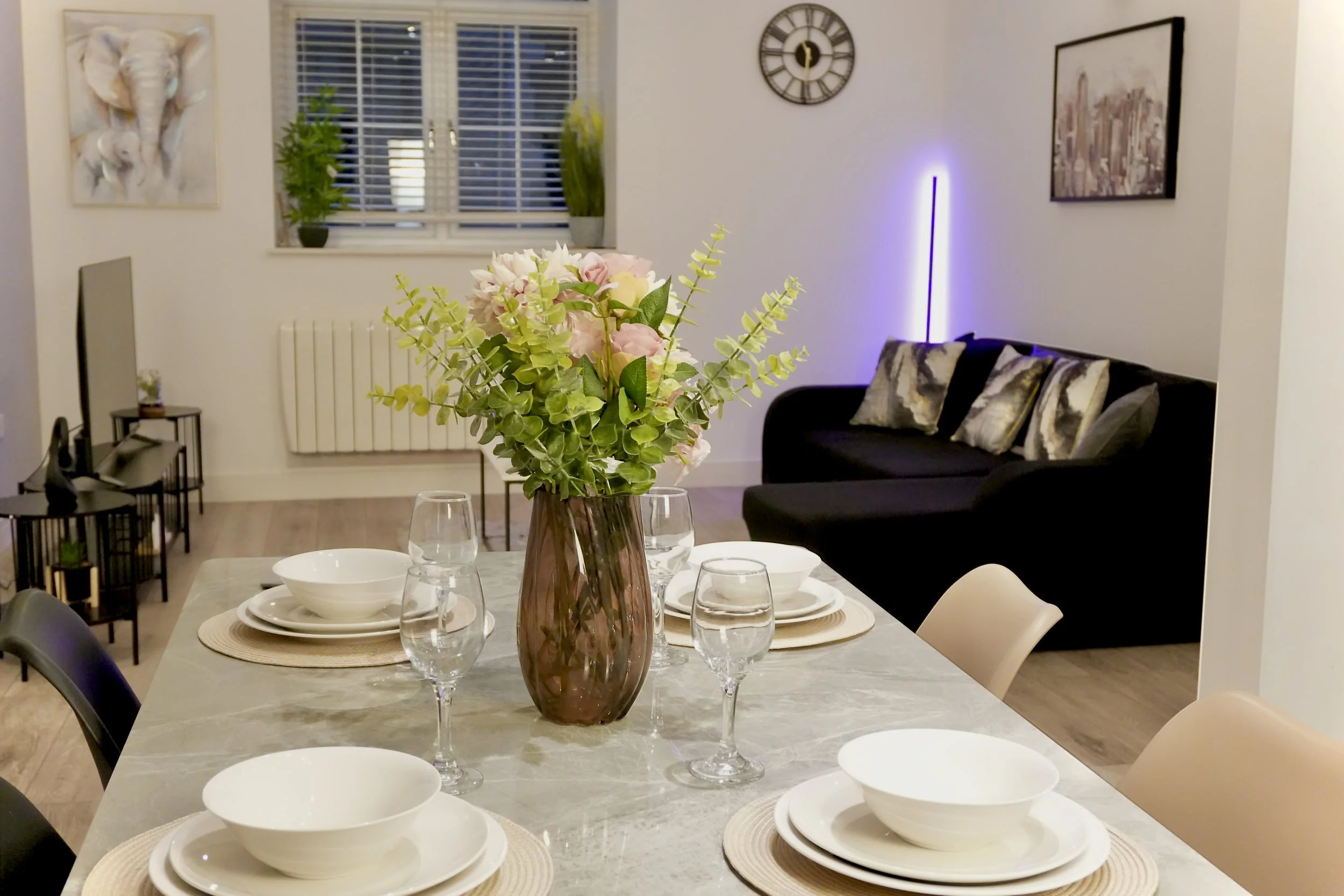 A dining table with a flower vase, four wine glasses, and four bowls set for a meal in a modern living room with a black sofa, wall art, and a clock.