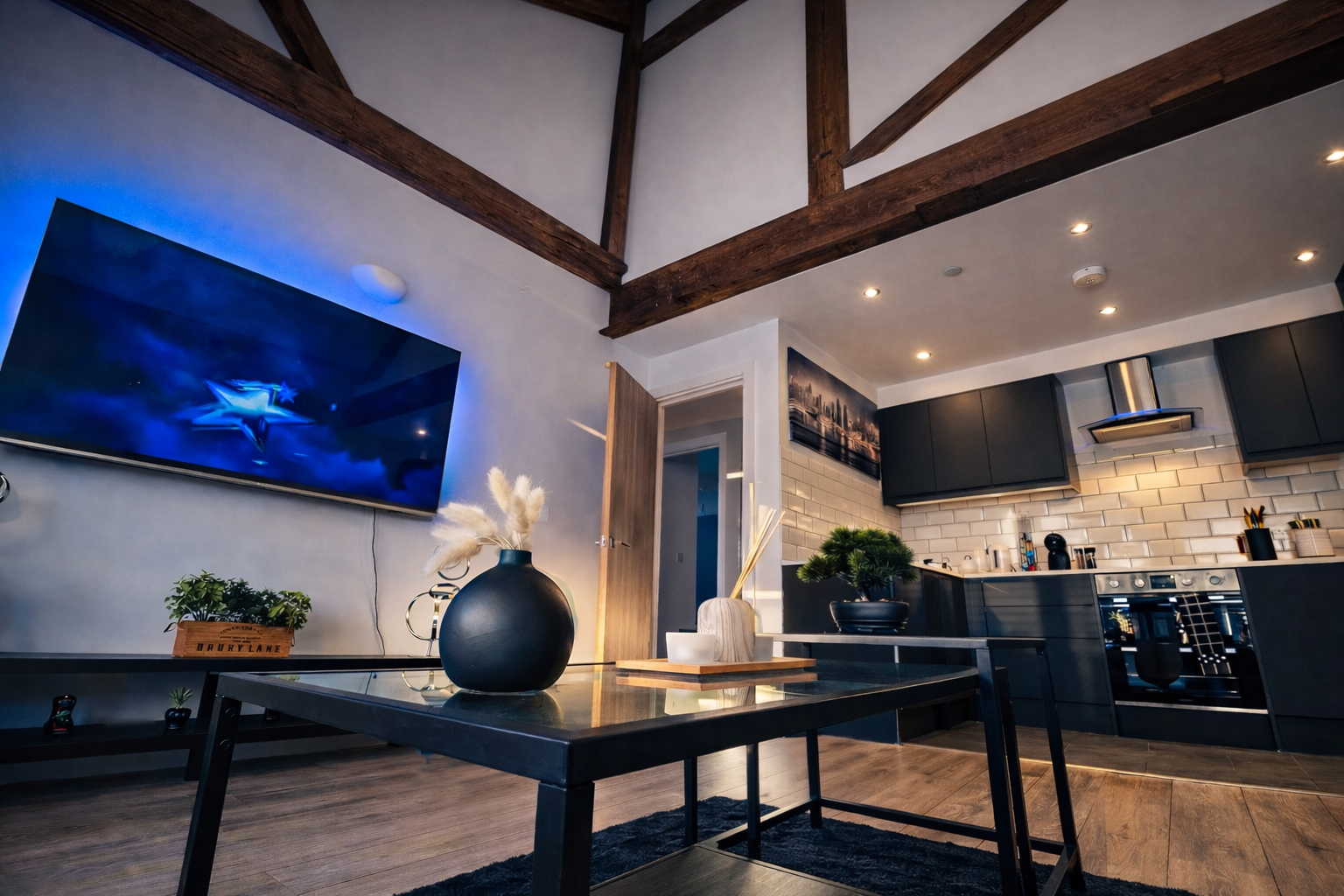 Modern kitchen and living room with exposed wooden beams, flat-screen TV, black cabinets, white subway tile backsplash, and decorative plants.