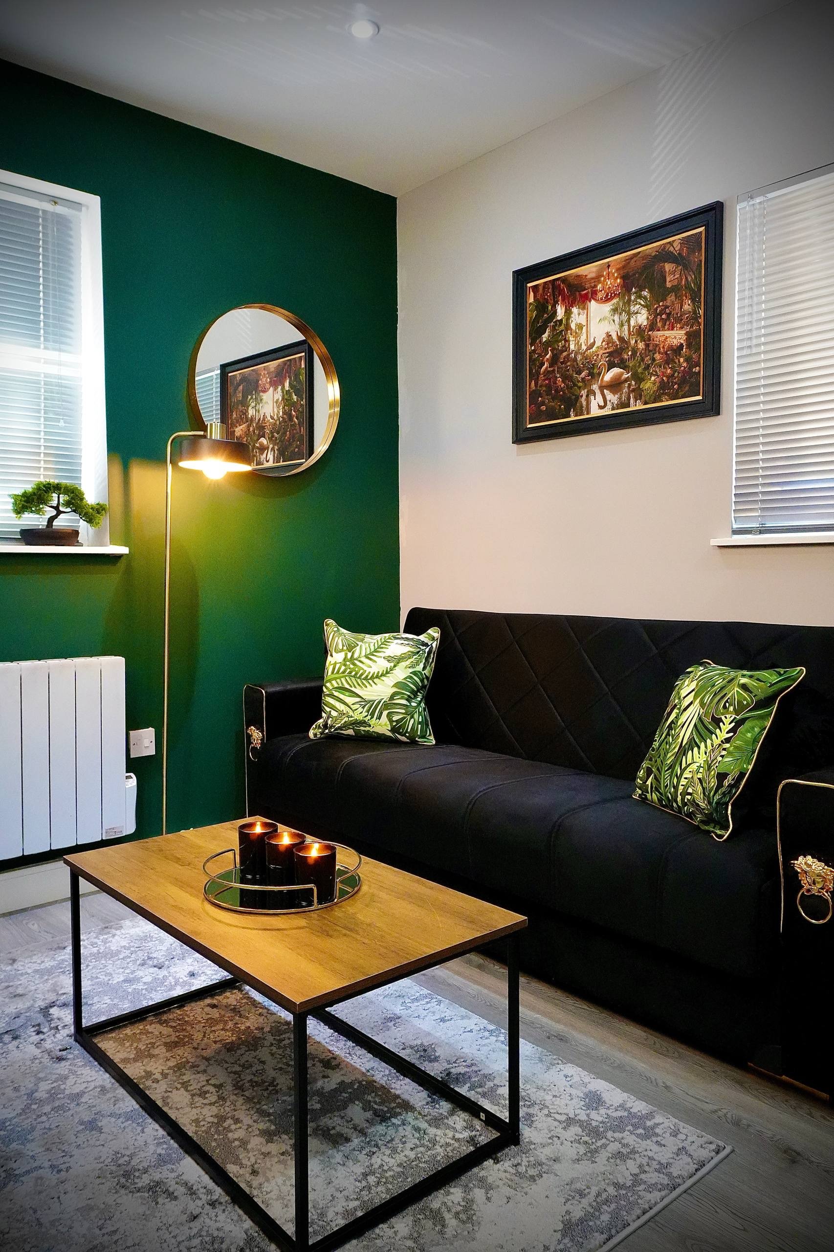 Modern living room with black sofa, white walls, wooden flooring, a wall clock, a window with blinds, potted plants, a TV on black stand, a small side table with decor, and a white coffee table on a rug.