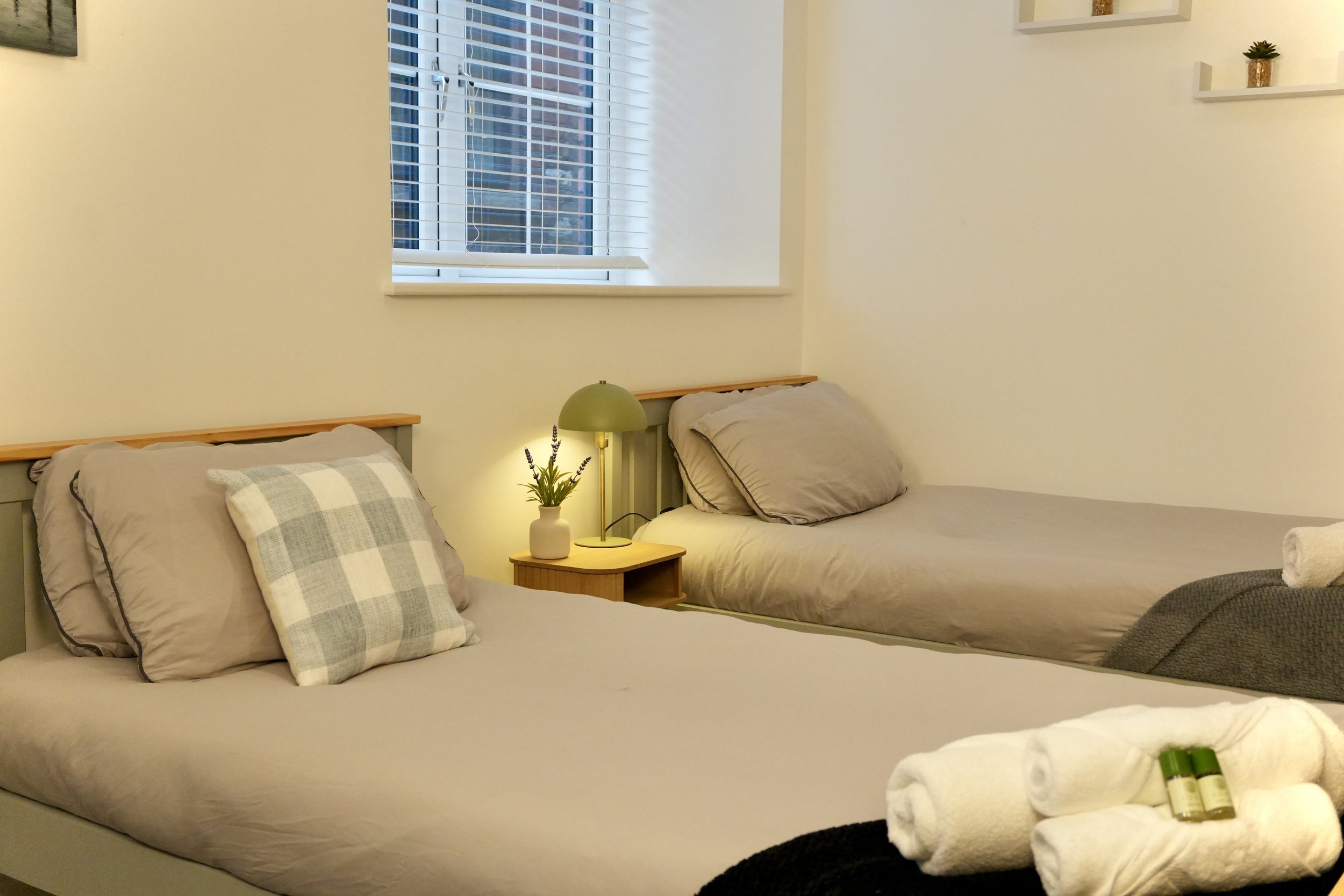 A cozy bedroom with two beds, a wooden nightstand, a small green table lamp, a vase with lavender flowers, and rolled towels with toiletries on the foreground. The room has beige walls, a window with blinds, and decorative shelves with small plants.