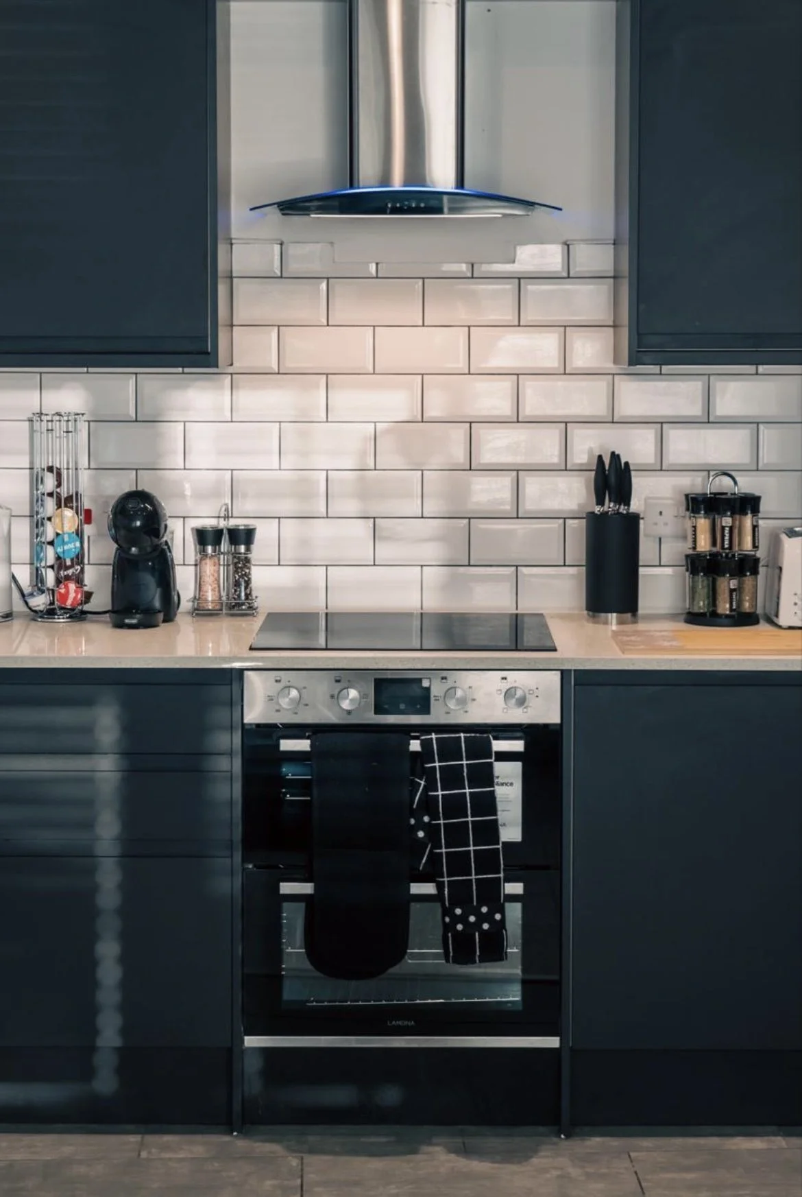 Modern kitchen with black cabinets, white tile backsplash, and stainless steel appliances, including an oven with towels hanging on it, and various kitchen items on the countertop.
