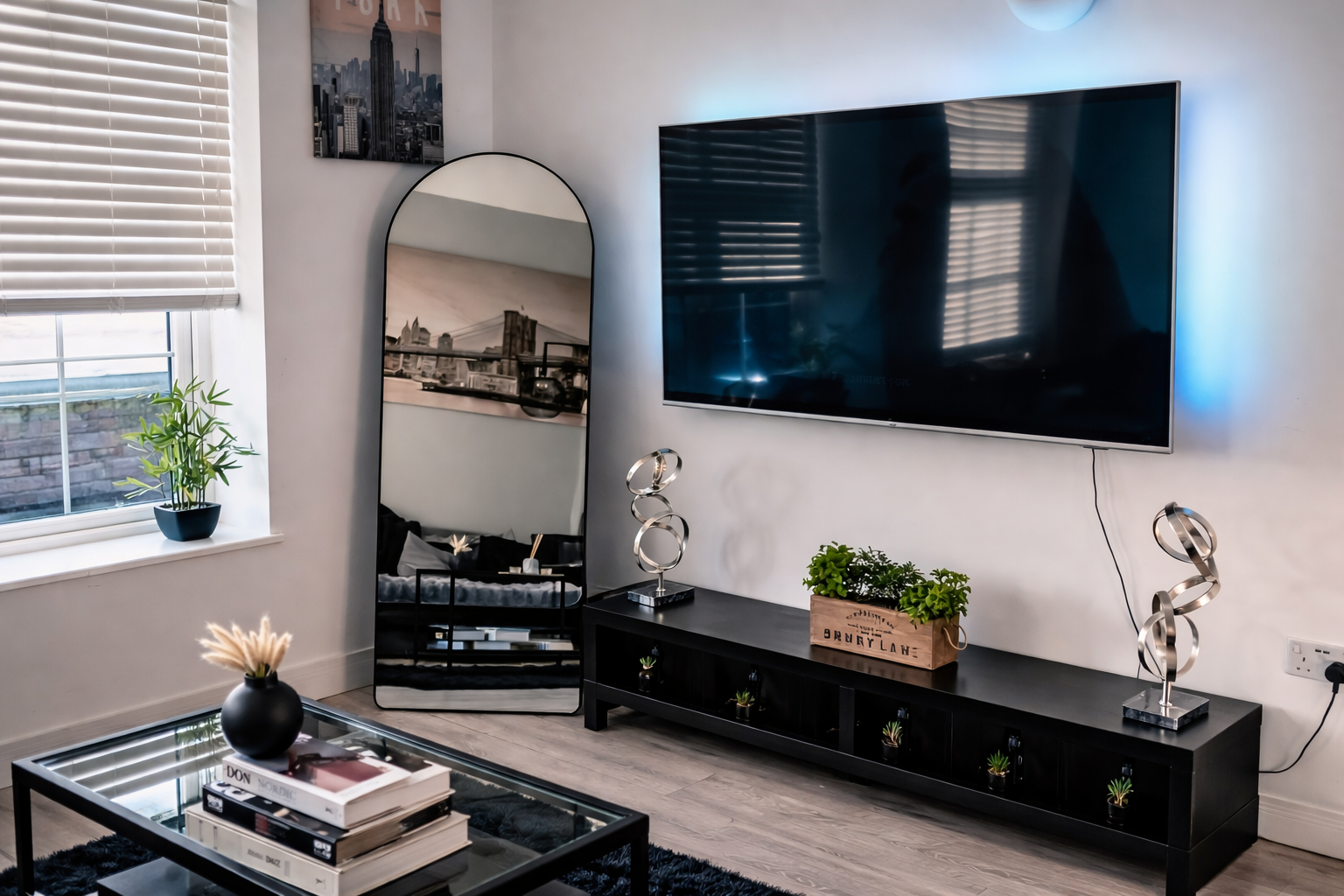 Living room with a large wall-mounted TV, a black console table with plants and decorative sculptures, a full-length mirror, a window with blinds, and a coffee table with books and a black vase with dried plants.