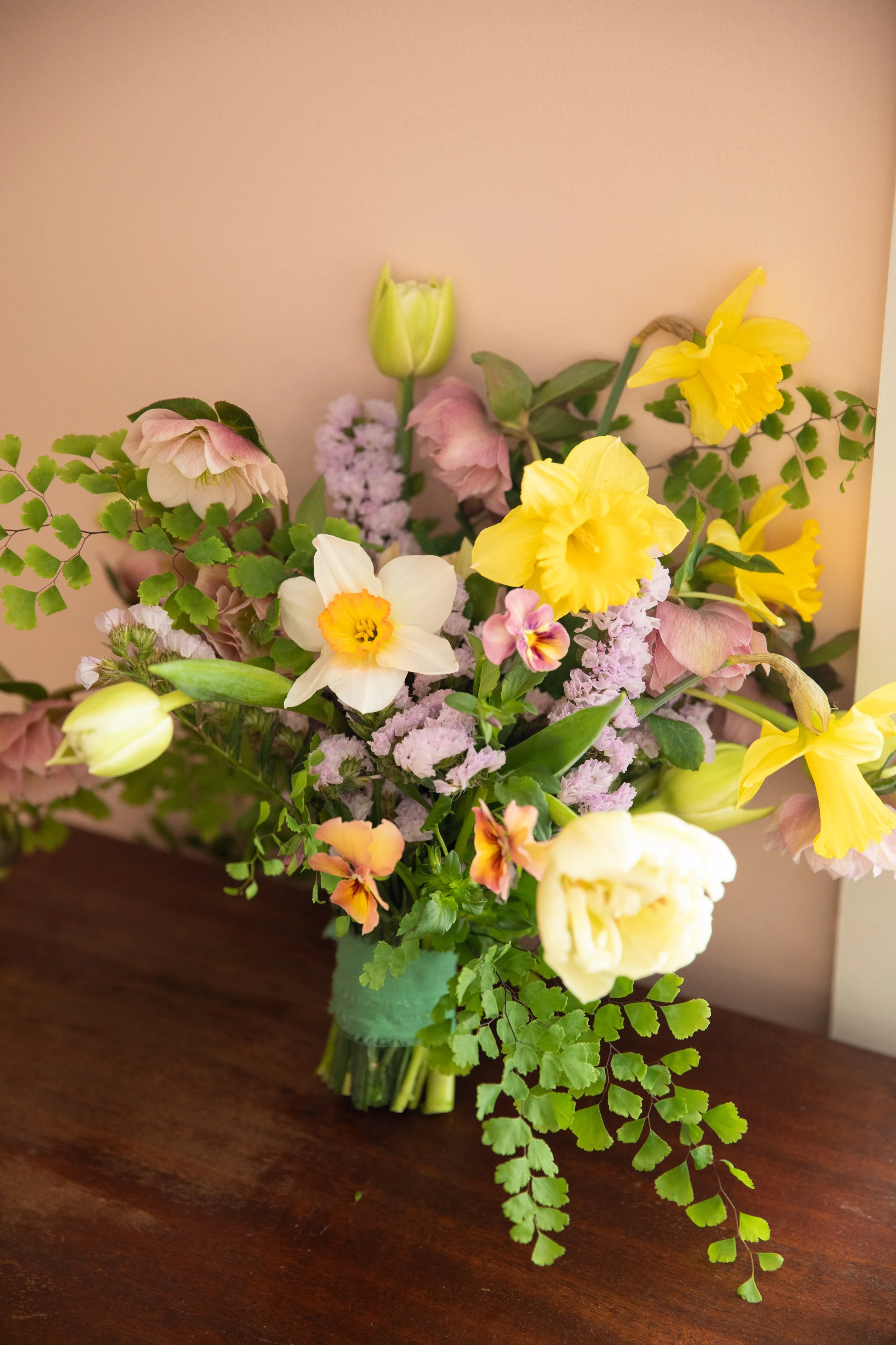 A colorful bouquet of various flowers including daffodils, tulips, and pansies in a green vase on a wooden surface.