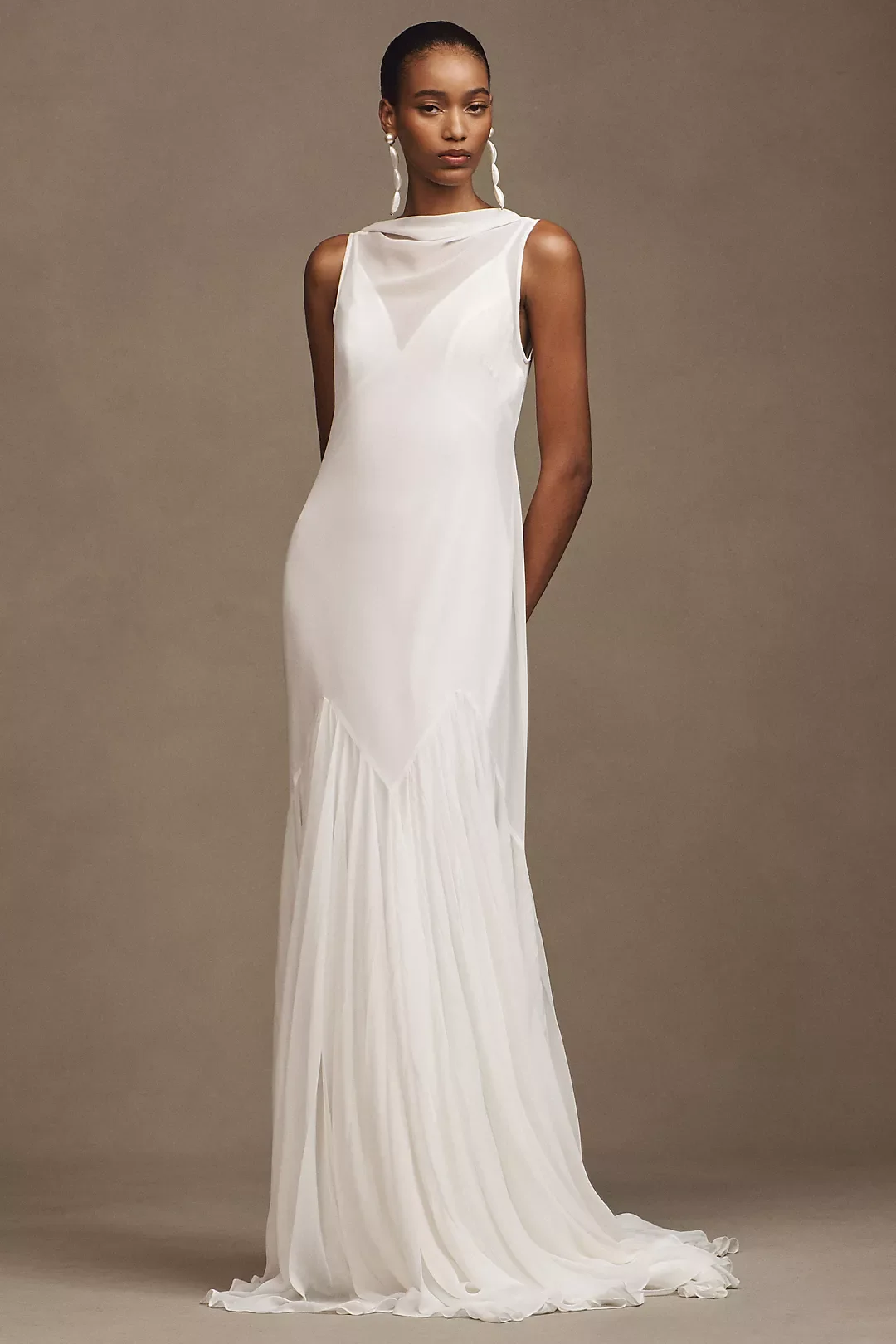 A woman in a white, sleeveless, flowing gown with a pleated skirt, standing against a neutral background, wearing statement earrings.