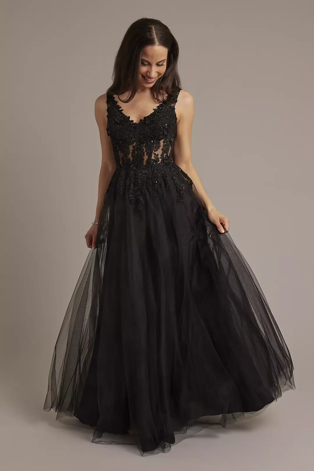 A woman in a black, floor-length, sleeveless, lace and tulle evening gown, smiling and looking down, holding the skirt of her dress.