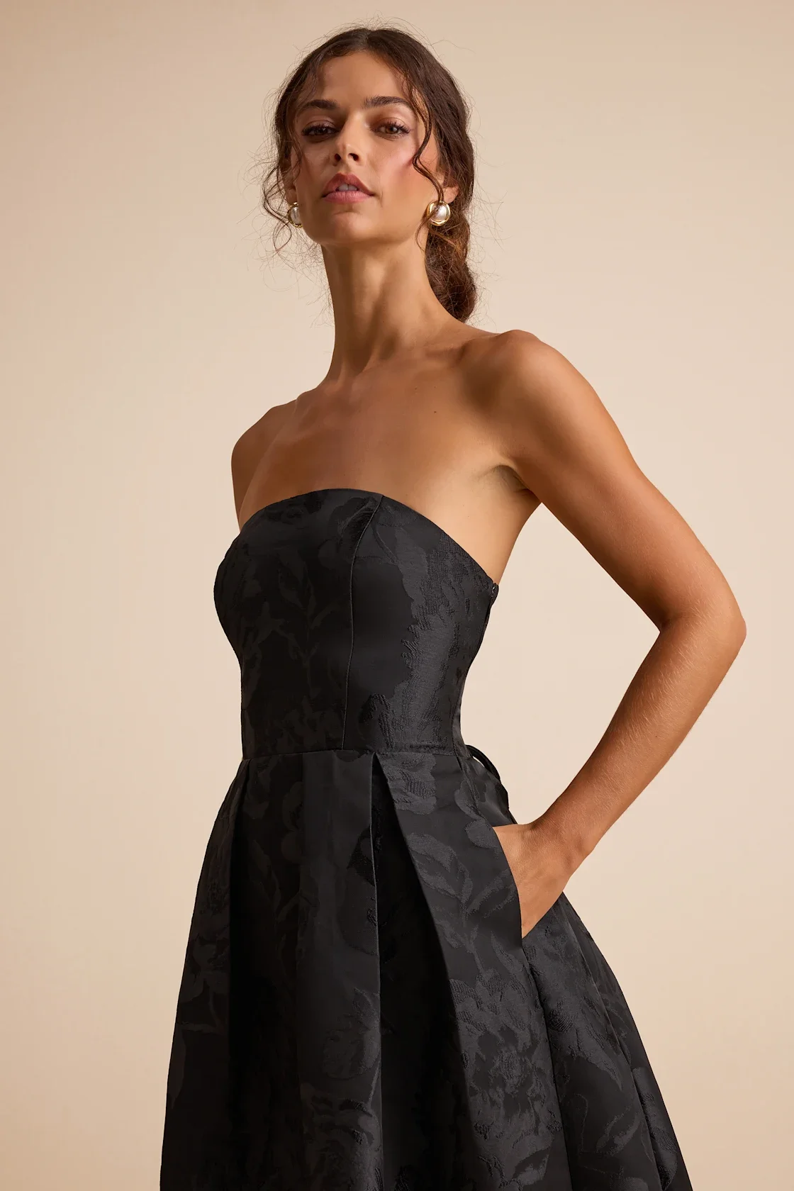 A woman with brown hair styled in loose waves, wearing pearl earrings and a strapless black dress with a subtle floral pattern, standing against a plain beige background.