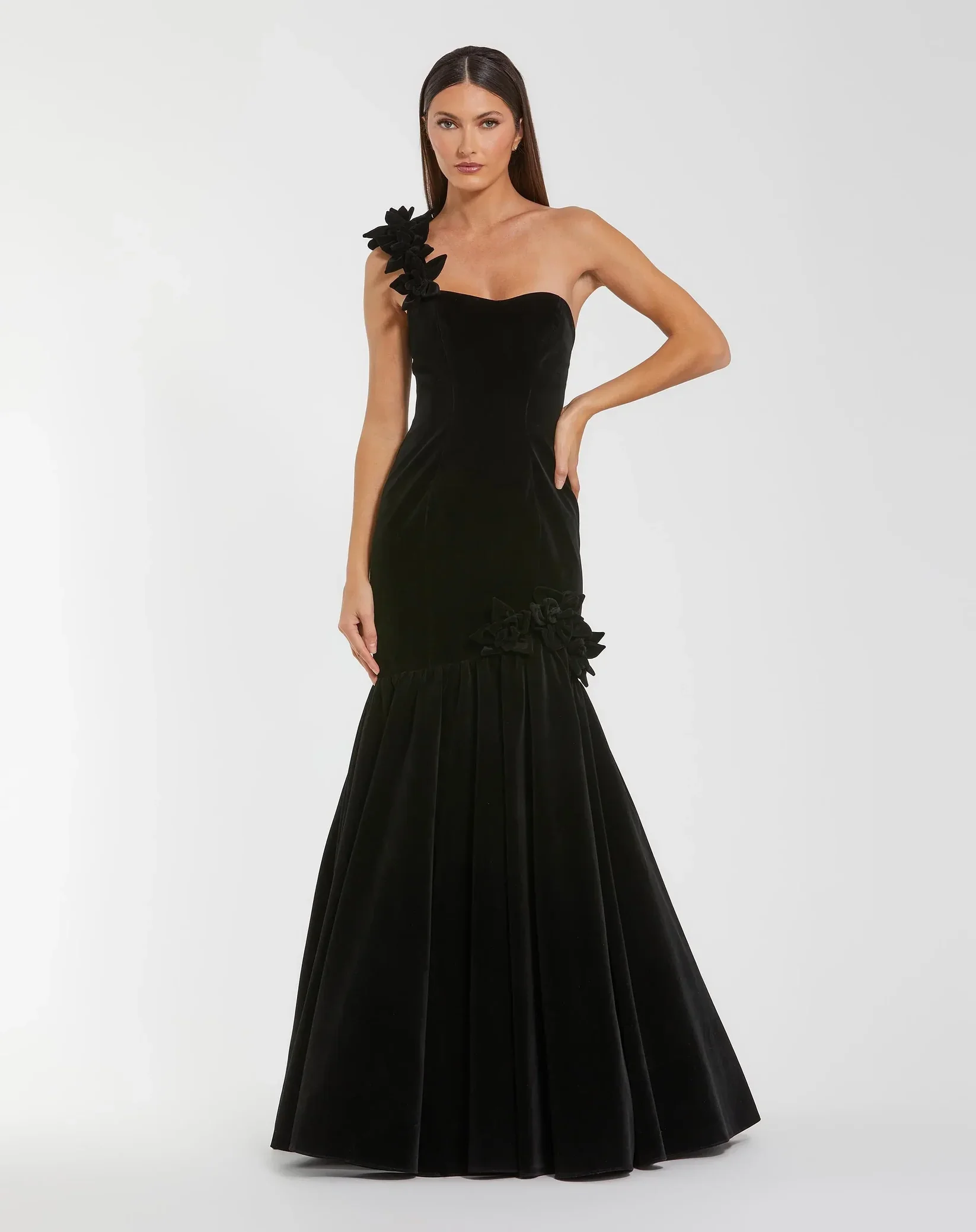 A woman with long dark hair wearing a strapless black velvet gown with floral embellishments on one shoulder and at the bottom of the skirt, standing against a plain white background.