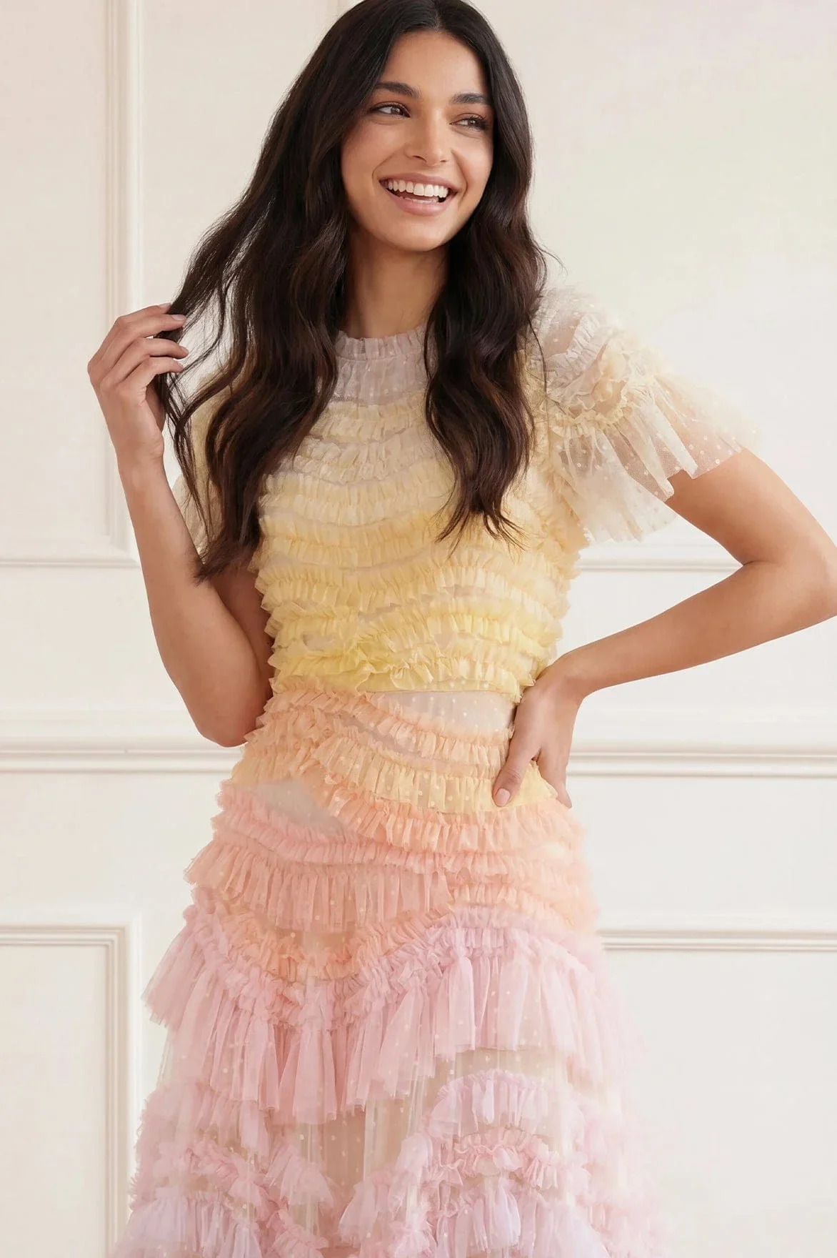 A woman with long dark hair smiling while wearing a pastel-colored tiered ruffled dress indoors.