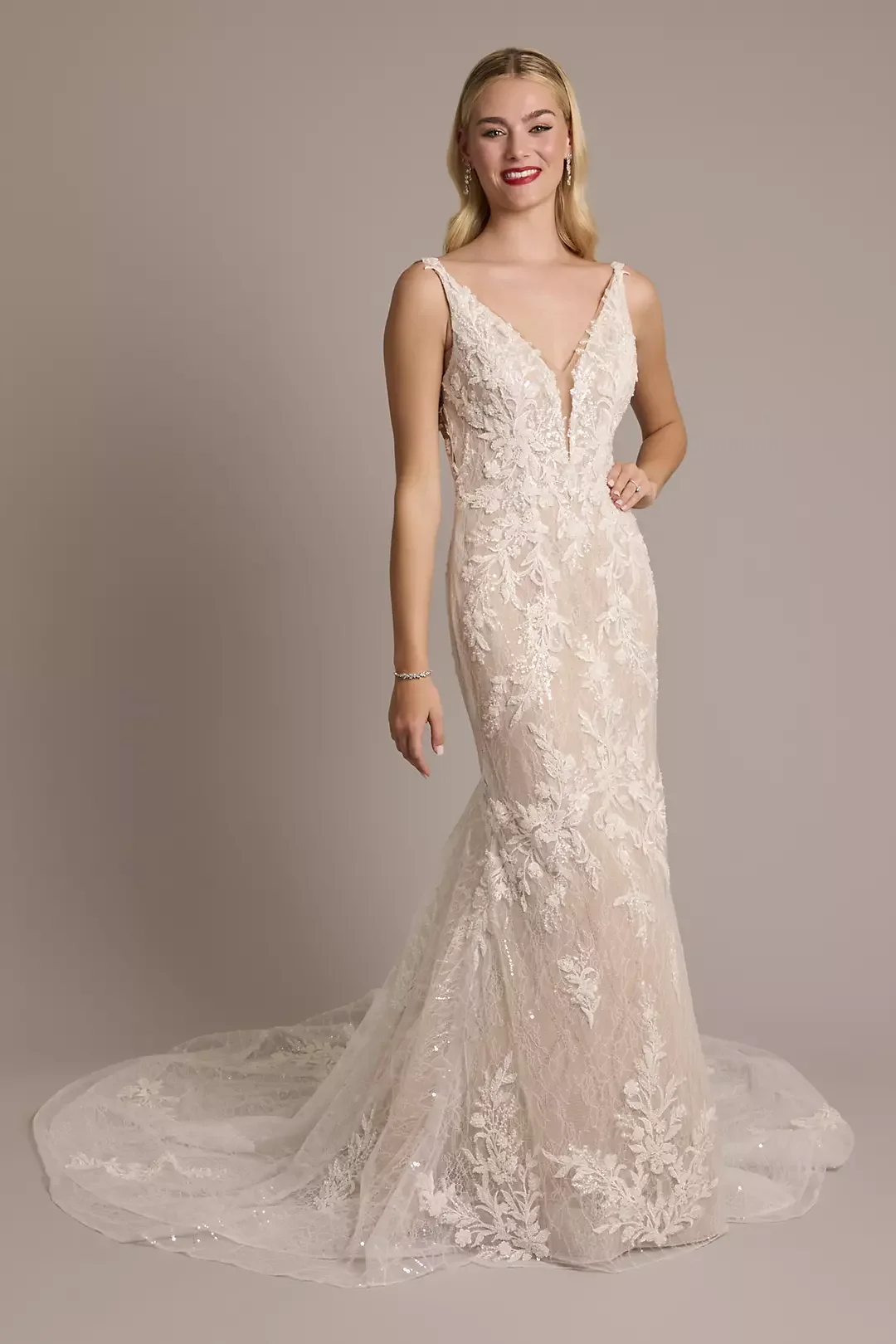 A woman in a lace wedding gown with a deep V neckline, fitted bodice, and train, standing against a plain background.