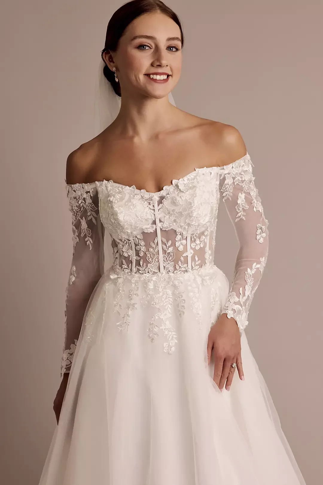 A smiling woman in a wedding dress with lace embroidery, off-the-shoulder neckline, and long sheer sleeves, standing against a plain background.